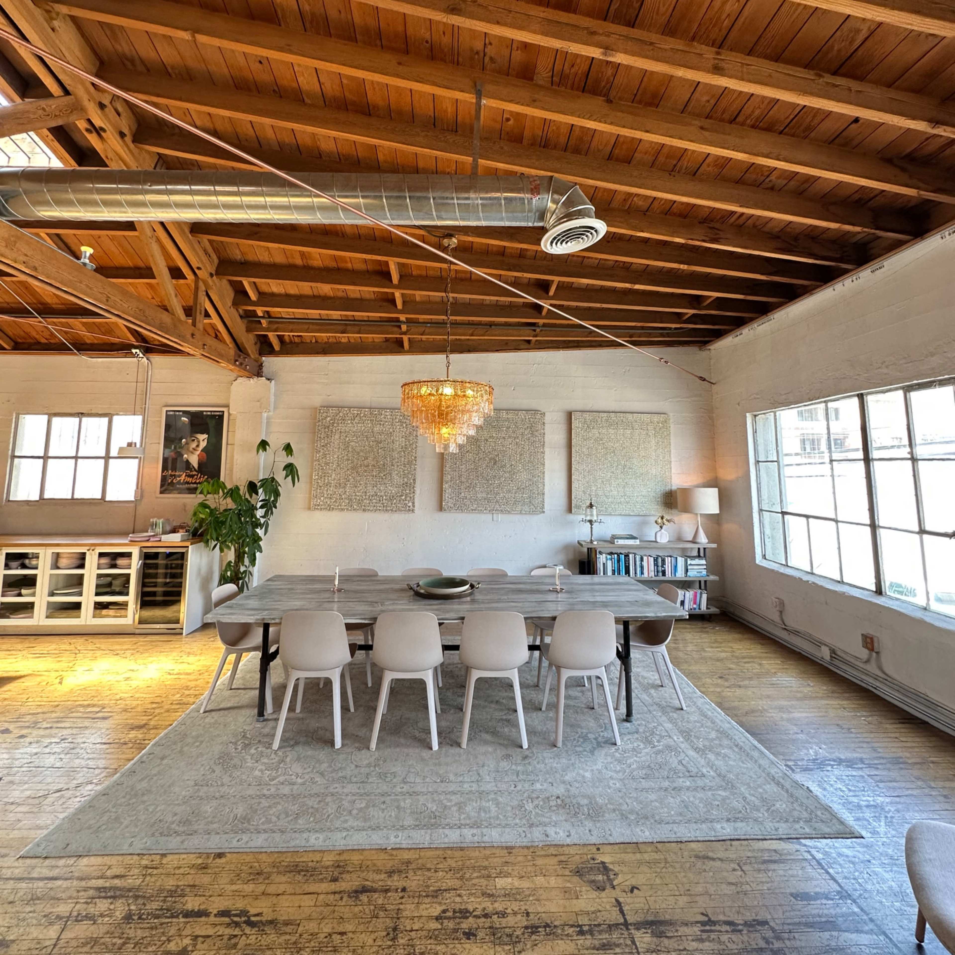A spacious dining area features a long table surrounded by white chairs, with a chandelier hanging above and large windows letting in natural light.