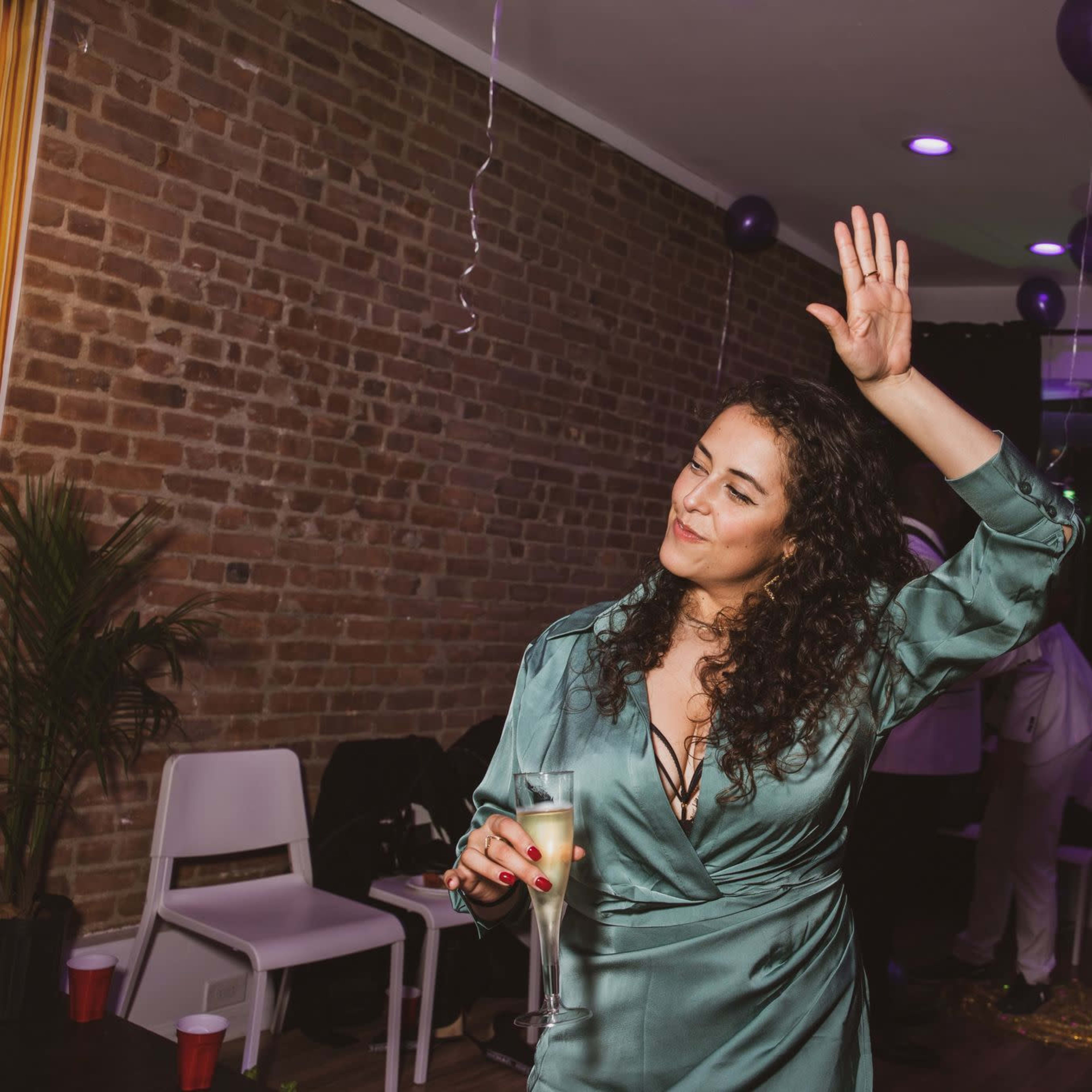 A woman in a green dress raises her hand while holding a drink at a festive indoor party with balloons and decorations.
