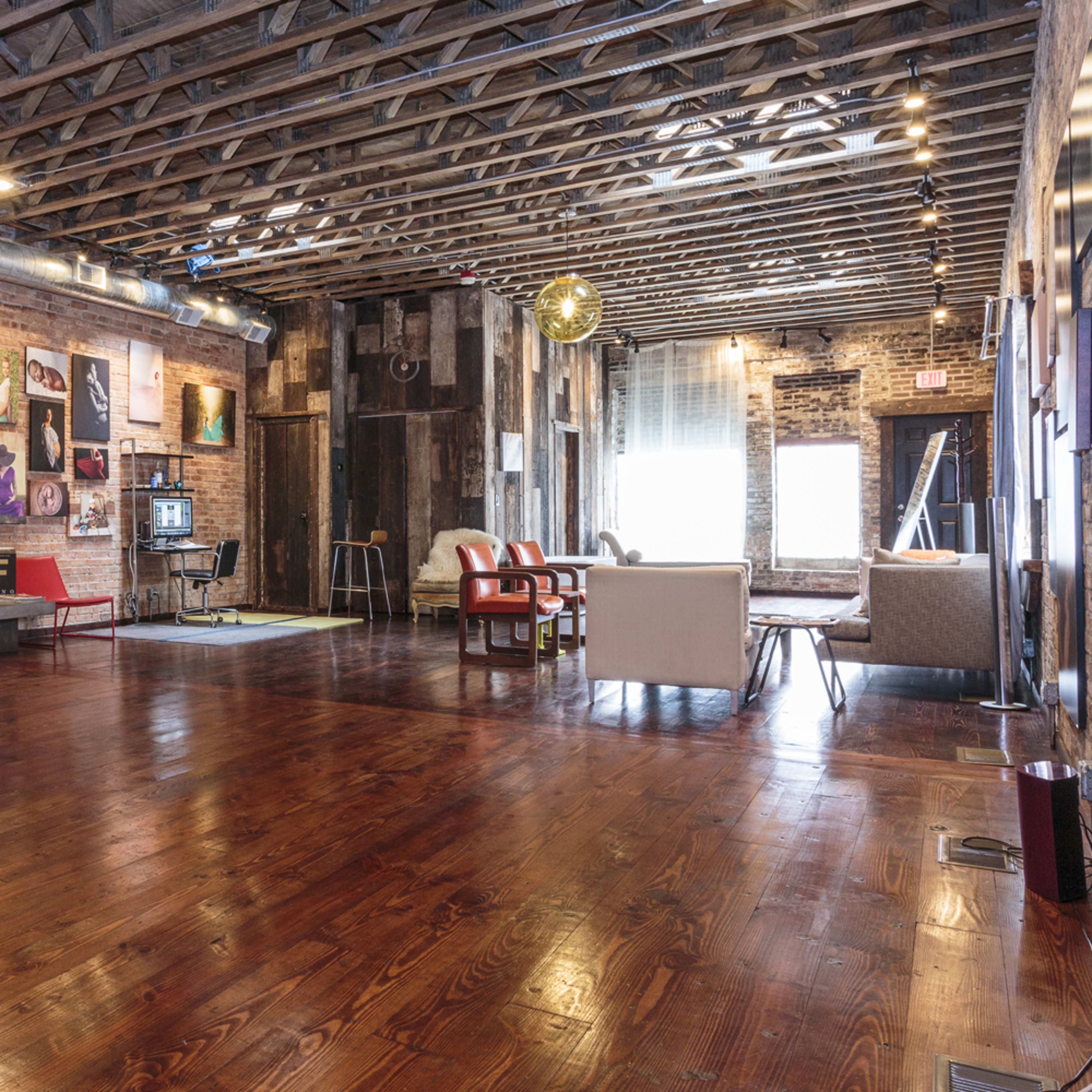 A spacious interior with exposed brick walls, wooden floors, and various seating arrangements, including red chairs and sofas, complemented by framed photographs on the walls.