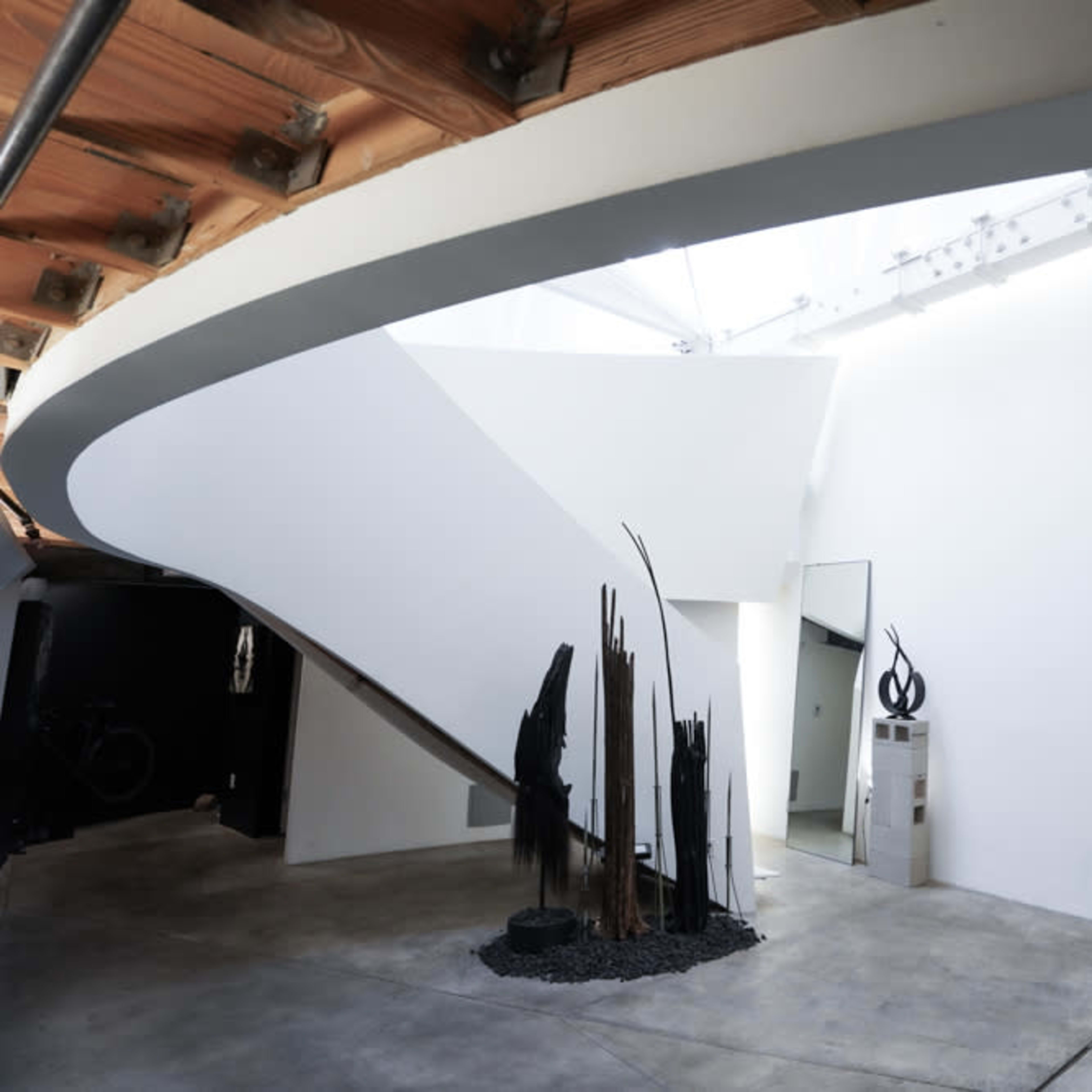 A modern interior space with a curved staircase leading to a minimalist hallway featuring sculptural elements and a light-colored wall.