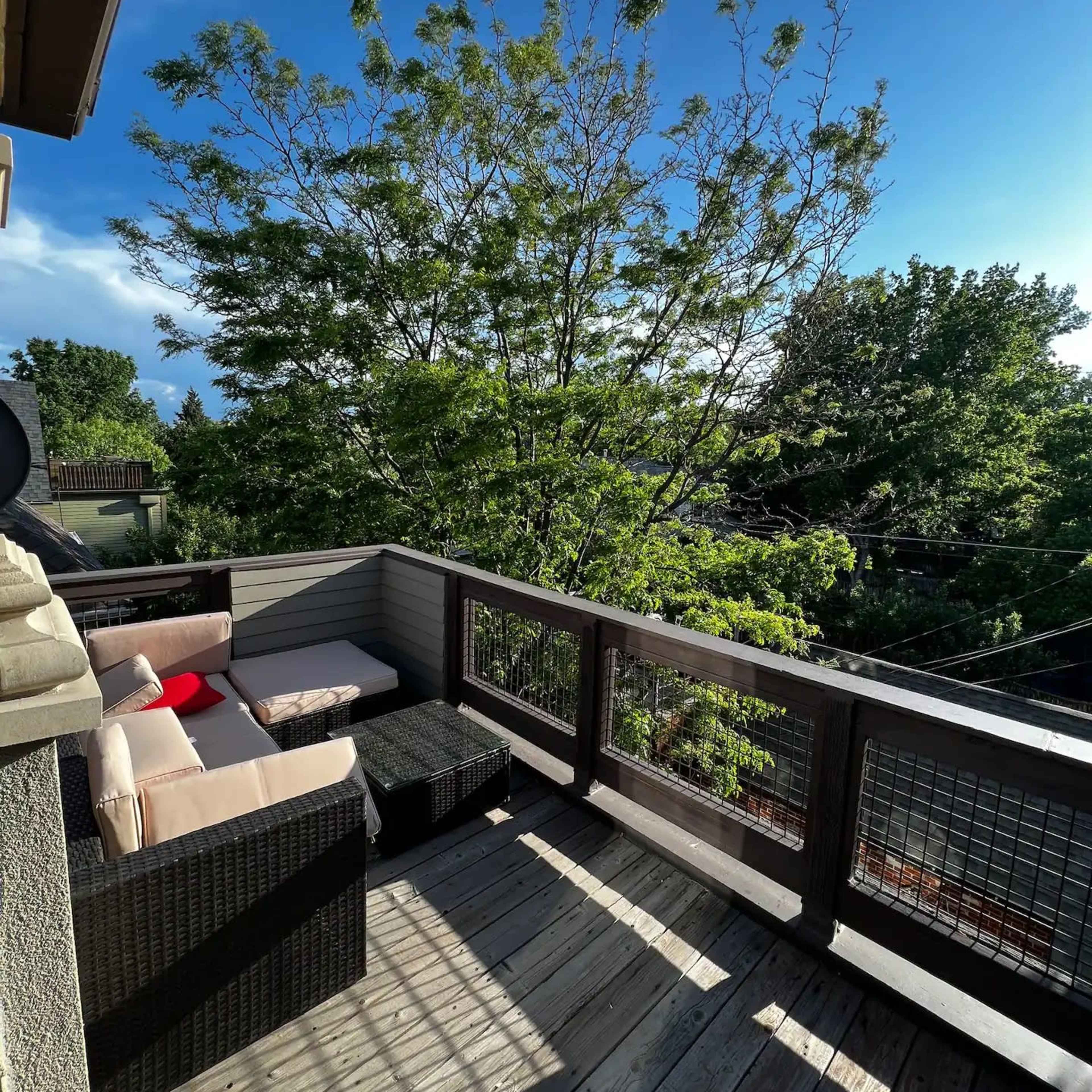 A wooden balcony features a seating area with a light-colored sofa and a black coffee table, surrounded by trees and blue sky.