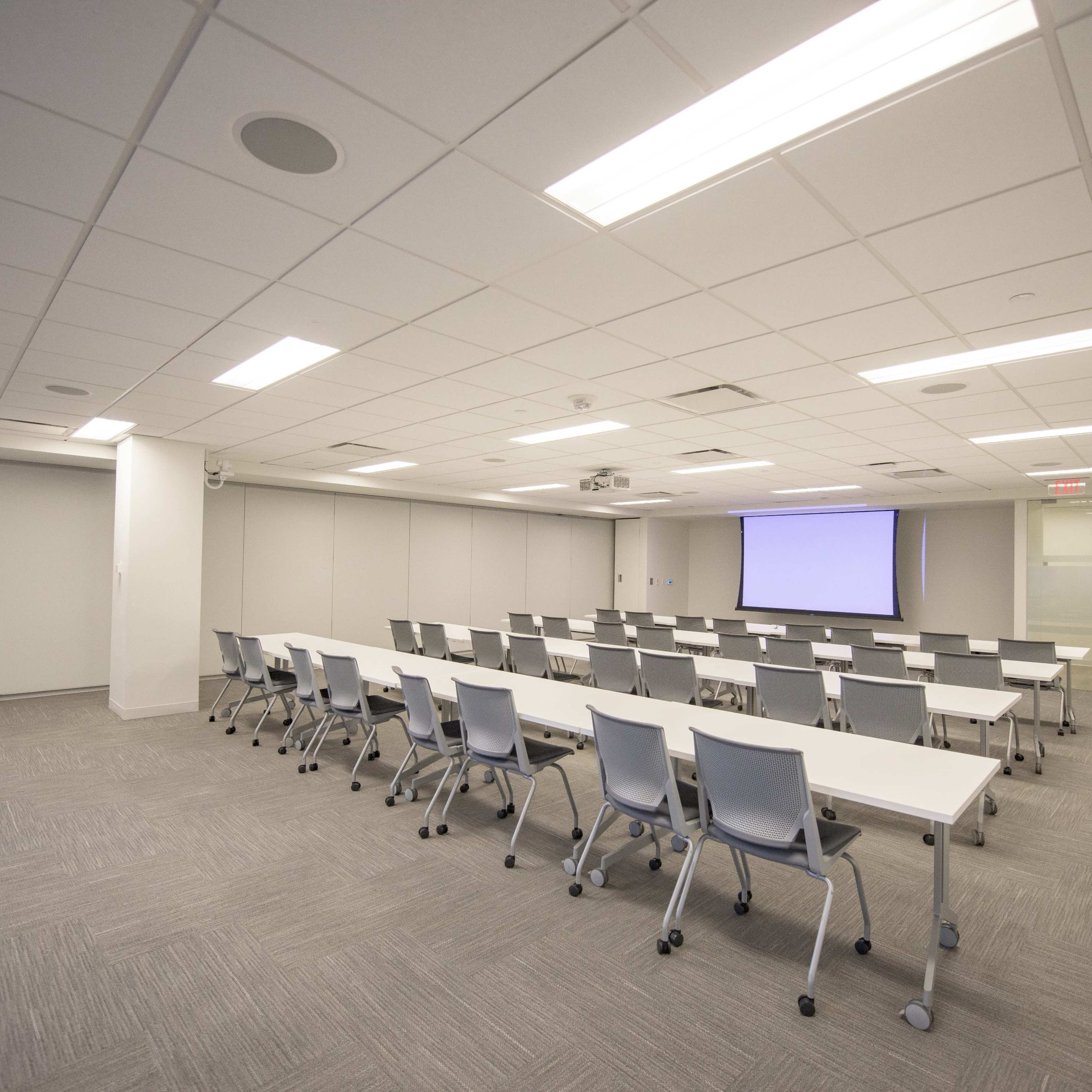 A conference room with rows of tables and chairs, equipped with a projector screen at one end.