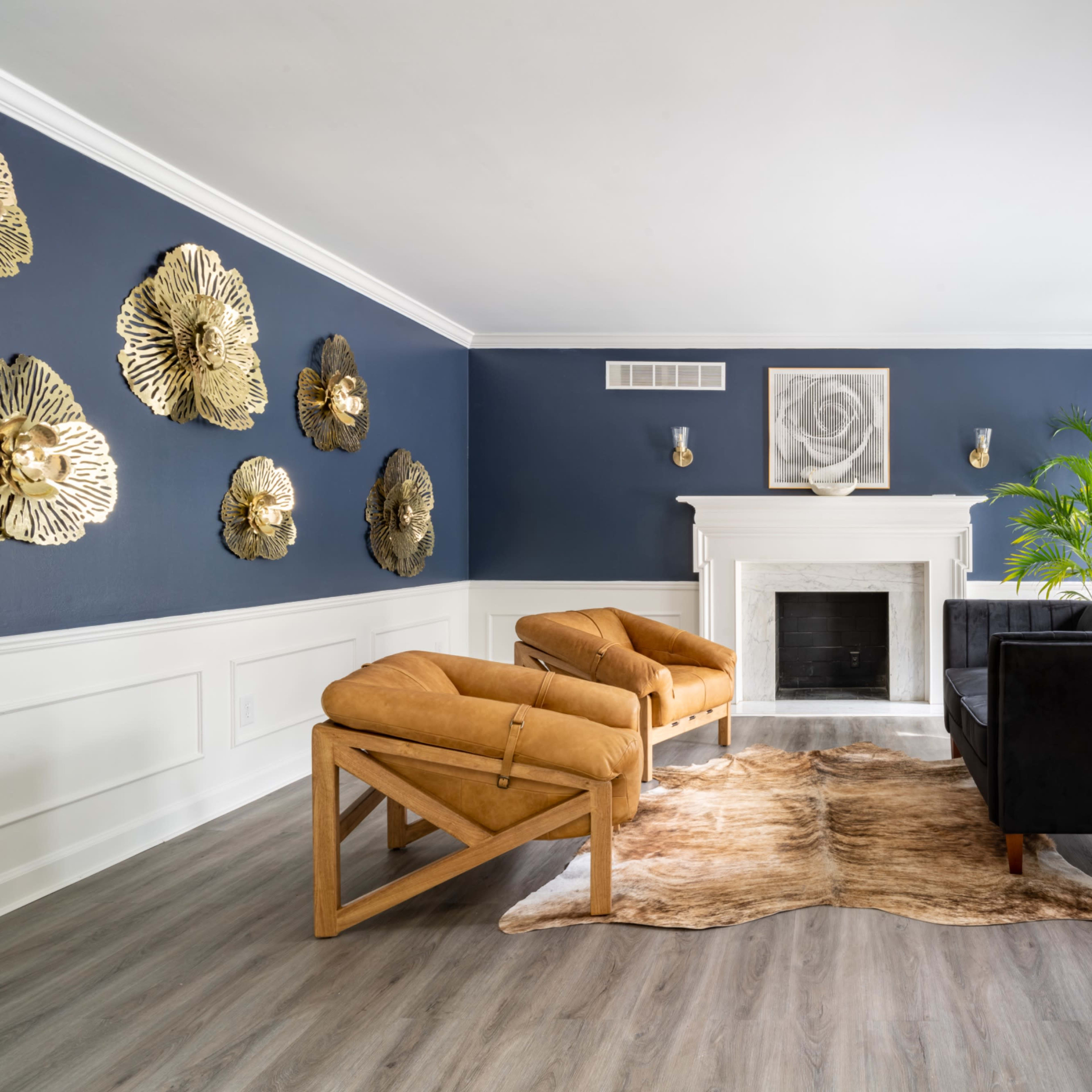 The room features a fireplace with a white mantel, two tan armchairs, a black sofa, a rug, and decorative wall art made of gold-colored flowers against dark blue walls.