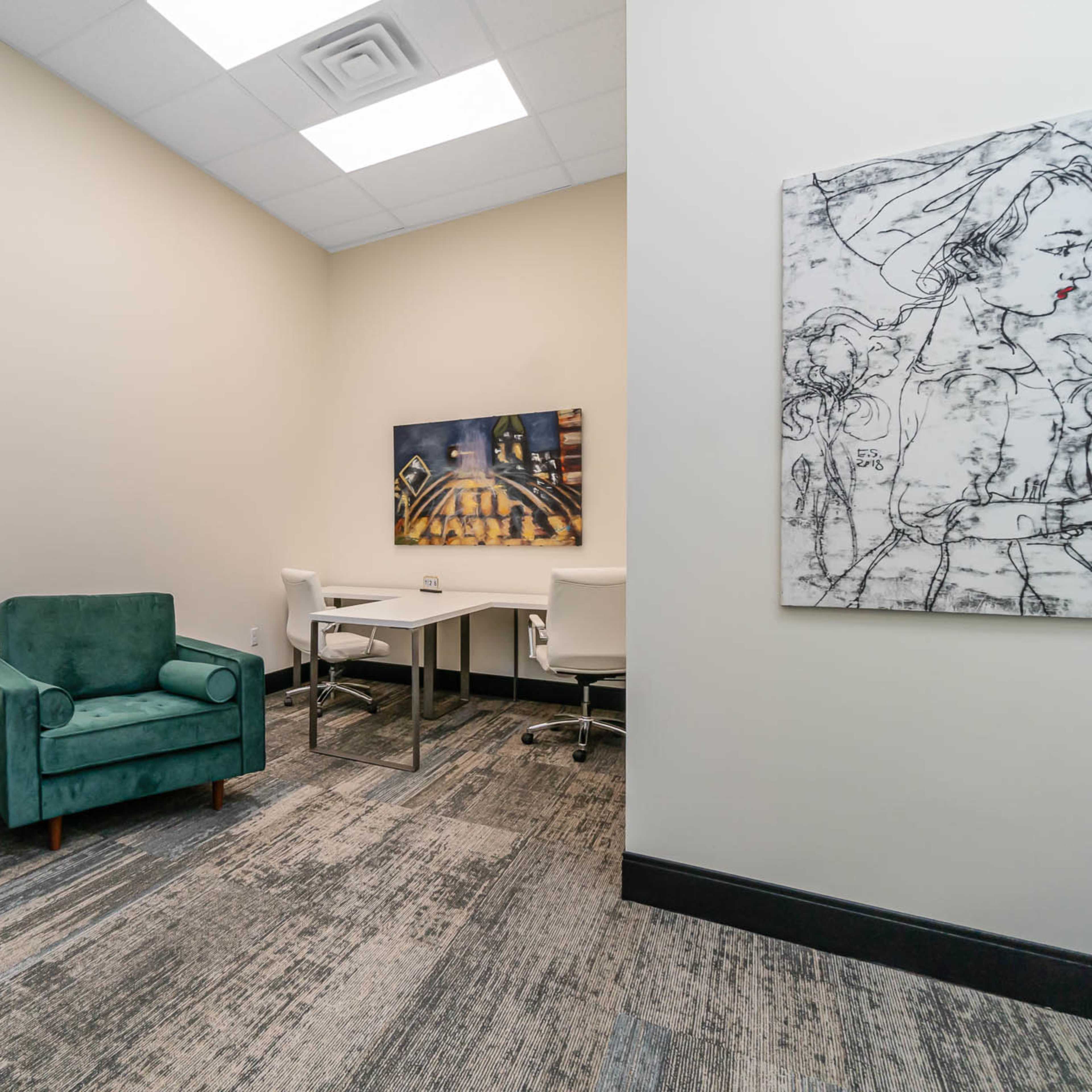 A small office space featuring two green armchairs, a modern desk with white chairs, and wall art on either side.
