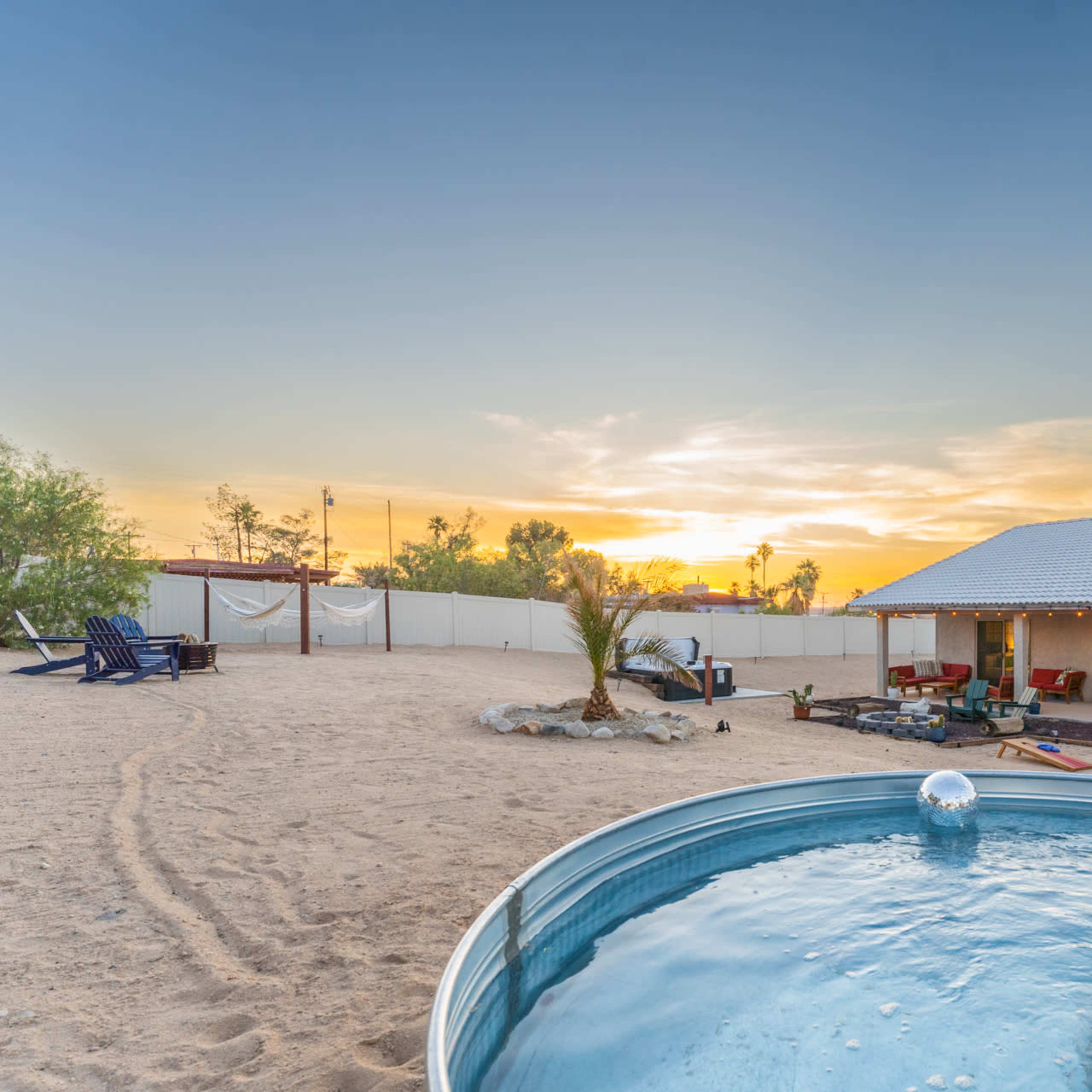 A backyard with a round pool, sandy ground, and a house with a patio overlooking a colorful sunset.