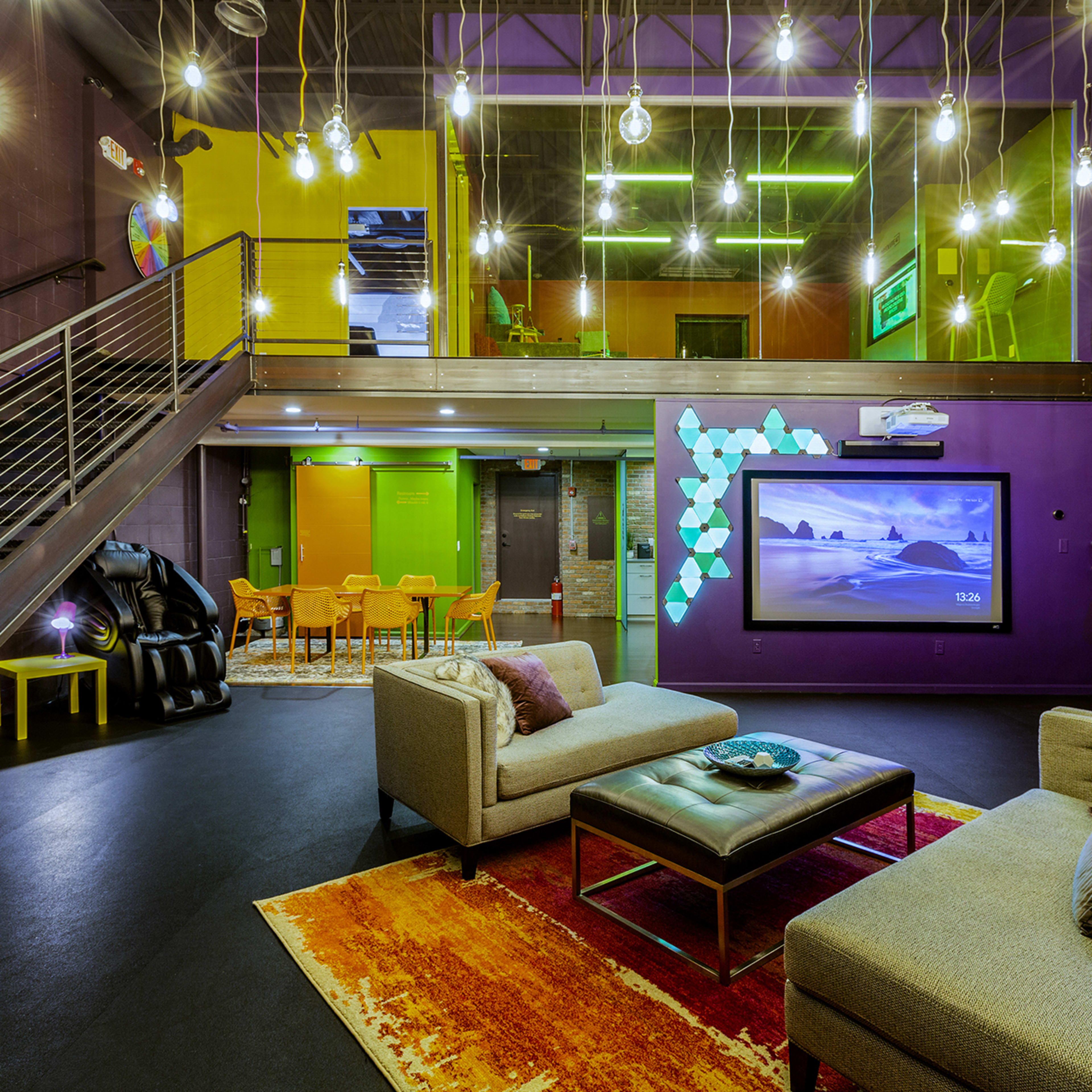 A modern lounge area with two sofas, a coffee table, and a large television, surrounded by bright lighting and vibrant colored walls.