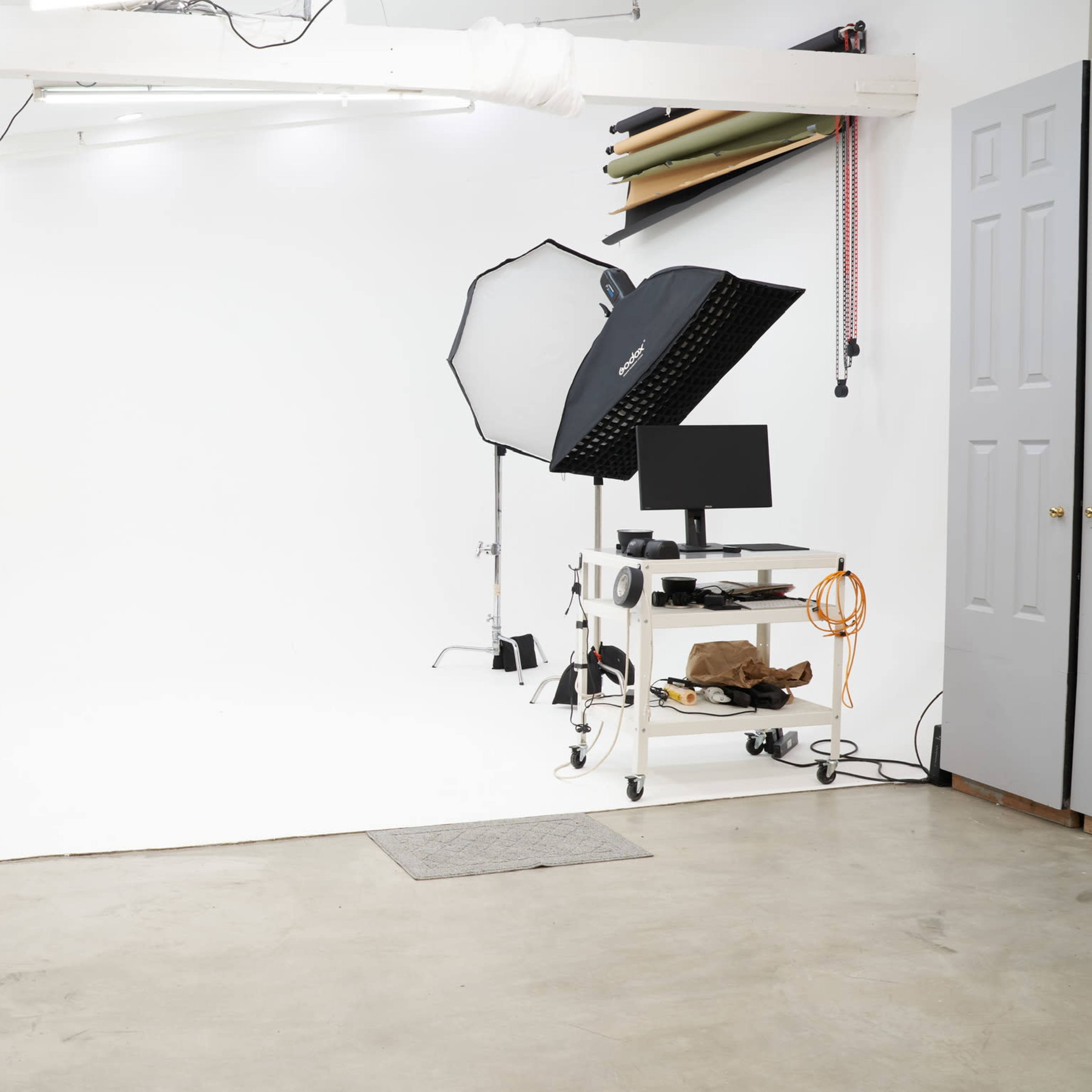 A blank photography studio with a white backdrop, lighting equipment, and a computer on a mobile cart.
