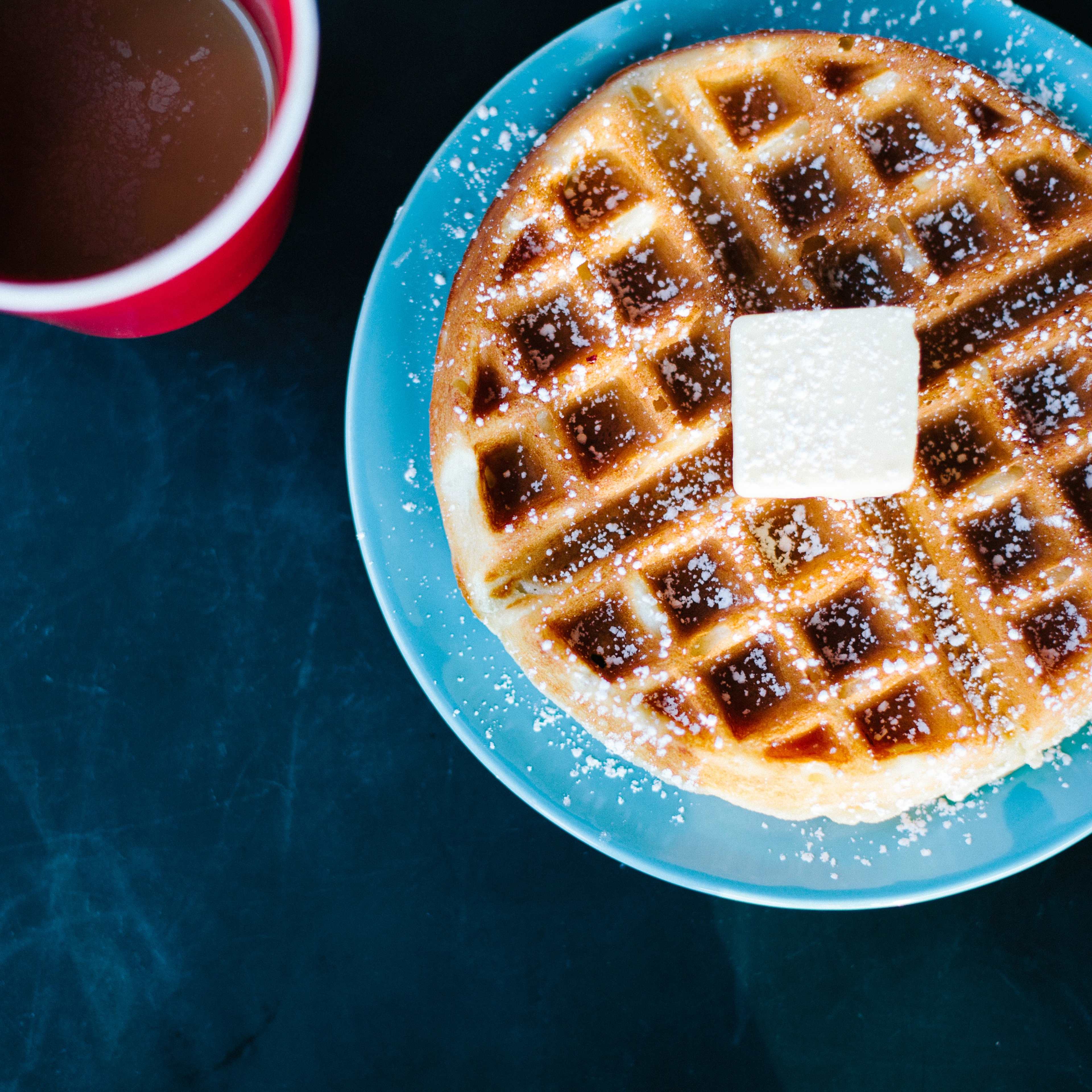 A golden waffle topped with a pat of butter sits on a blue plate next to a red cup filled with coffee.