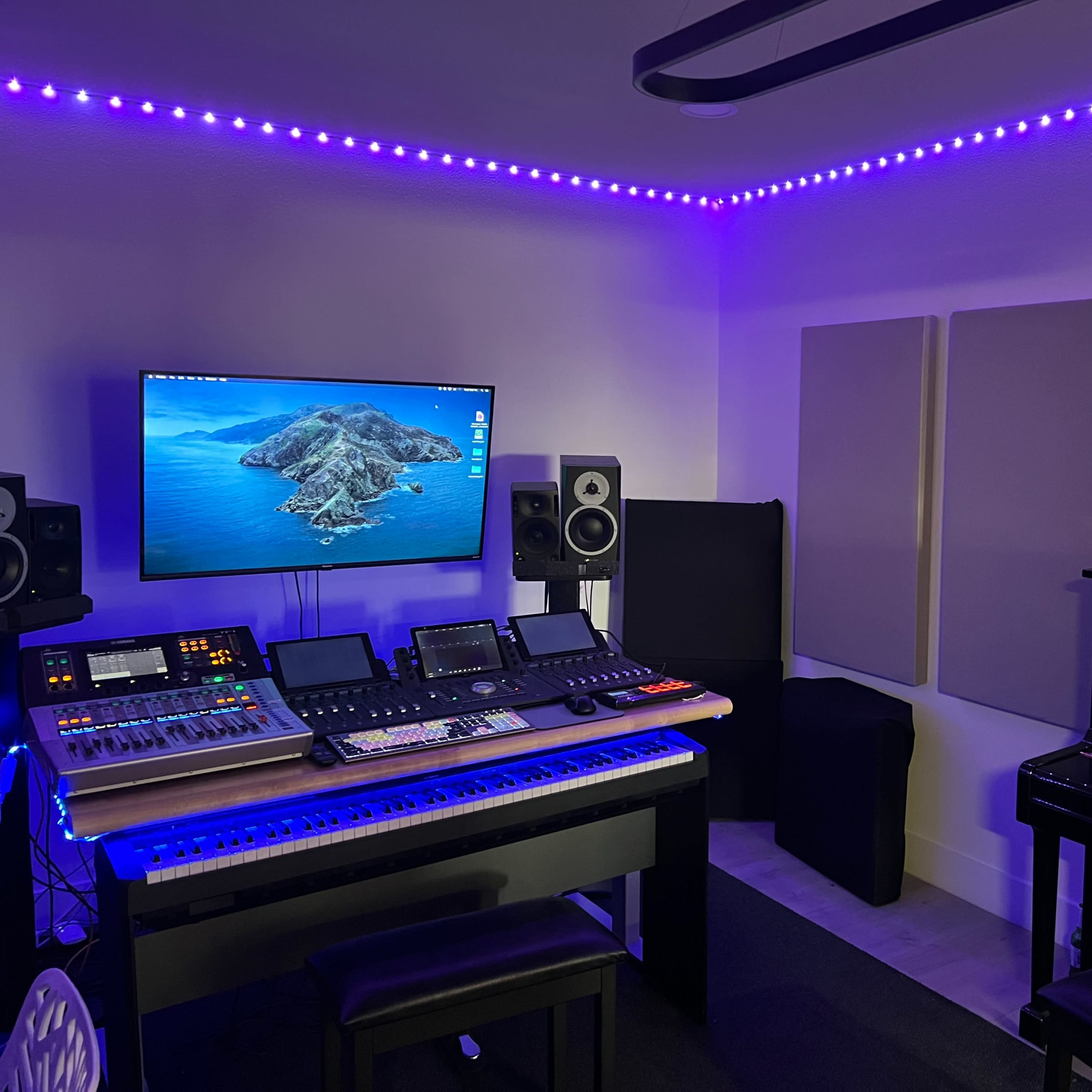 A music studio with a black piano, a keyboard setup, a computer monitor displaying a landscape, and sound equipment, all illuminated by purple LED lights.
