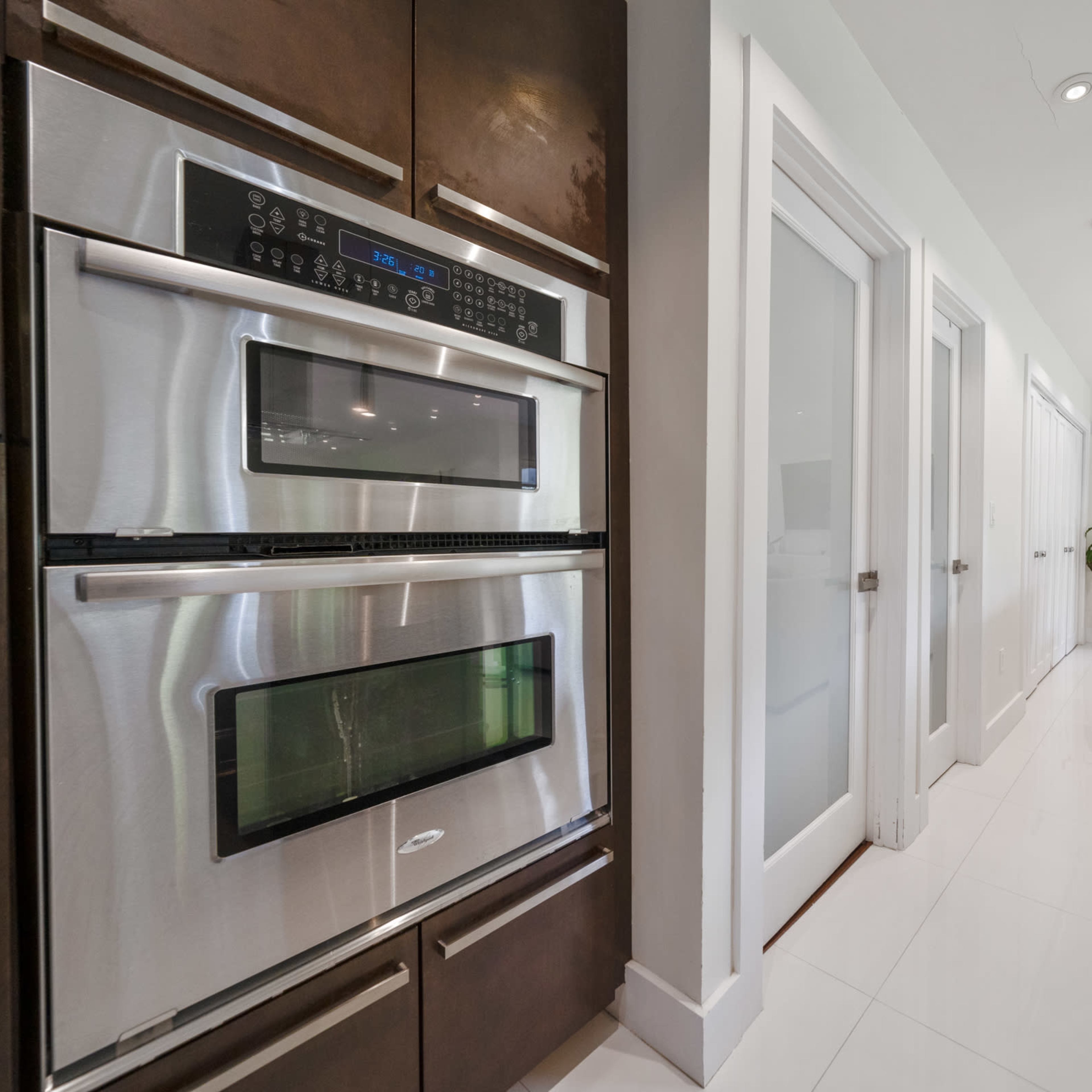 A modern kitchen area featuring stainless steel double ovens, a long white countertop, and a view into a bright living space with a large television.