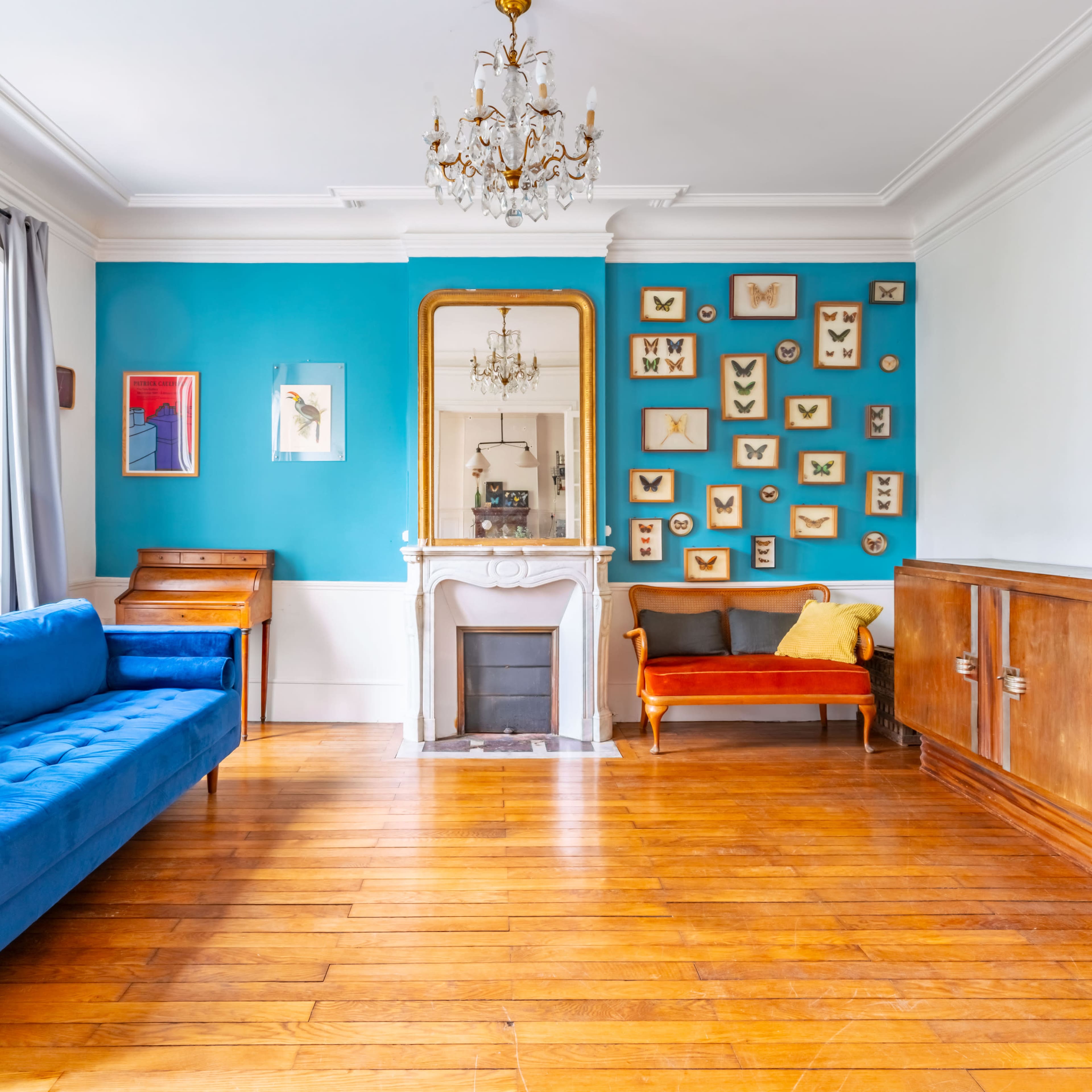 The living room features a blue sofa, a vintage armchair, a white fireplace, and a wall adorned with framed butterfly displays against a teal backdrop.