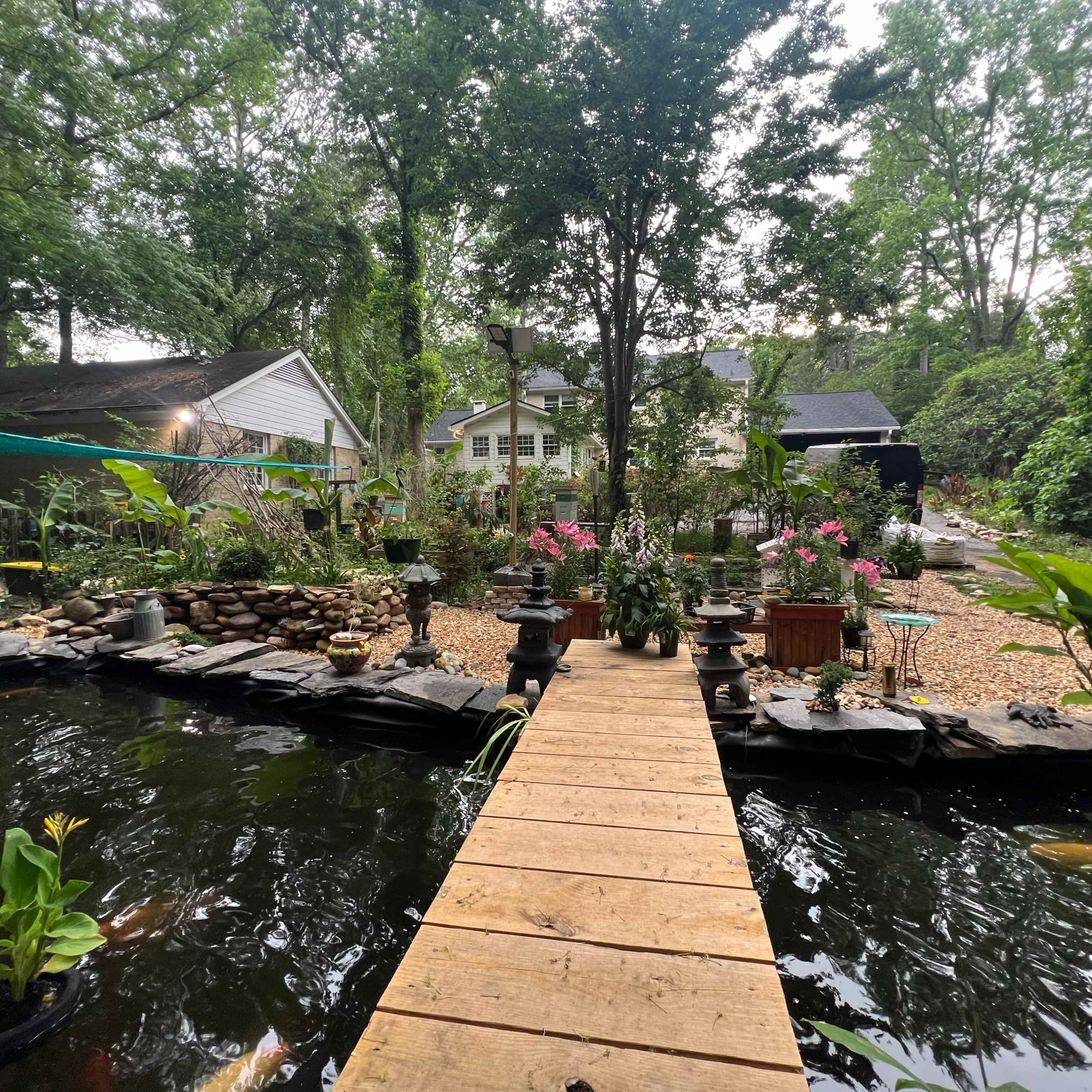 A wooden bridge crosses a pond surrounded by plants and flower beds in a garden setting.