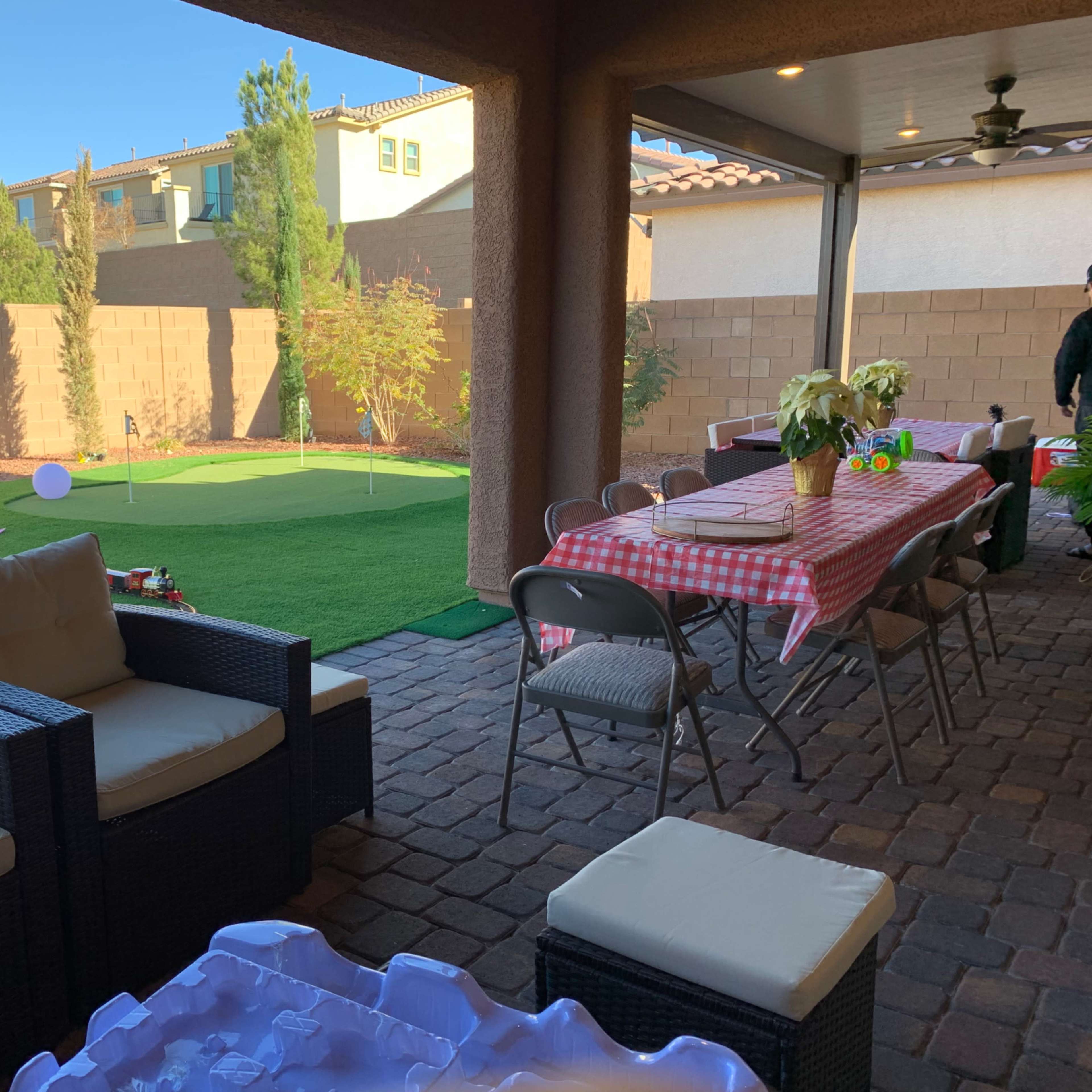 A backyard patio area with seating, a long table set for dining, and a green lawn featuring a putting green in the background.
