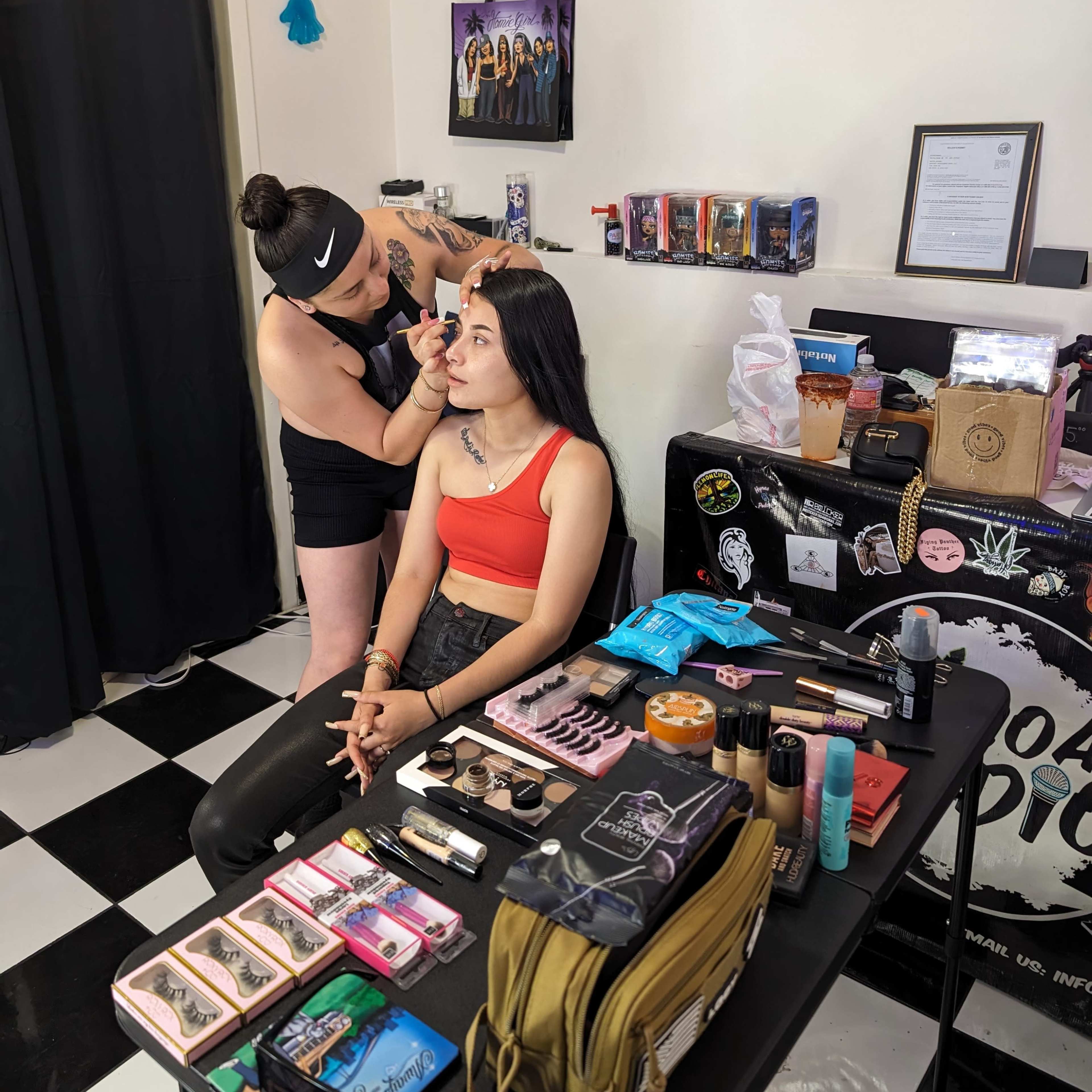 A makeup artist applies makeup to a client at a table filled with various cosmetics in a room with black and white checkered flooring.