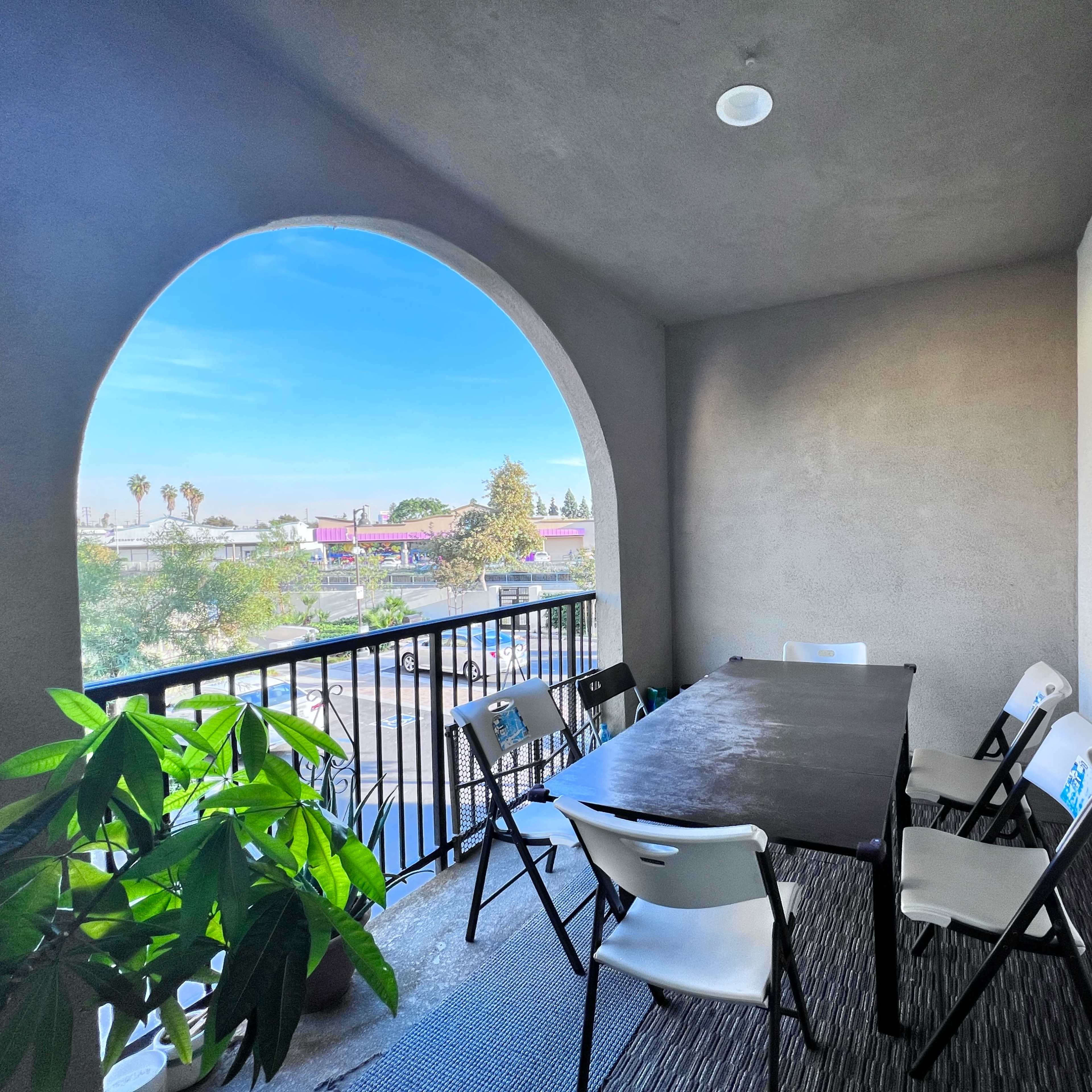 A covered balcony features a rectangular table surrounded by six folding chairs, with a view of palm trees and buildings outside.
