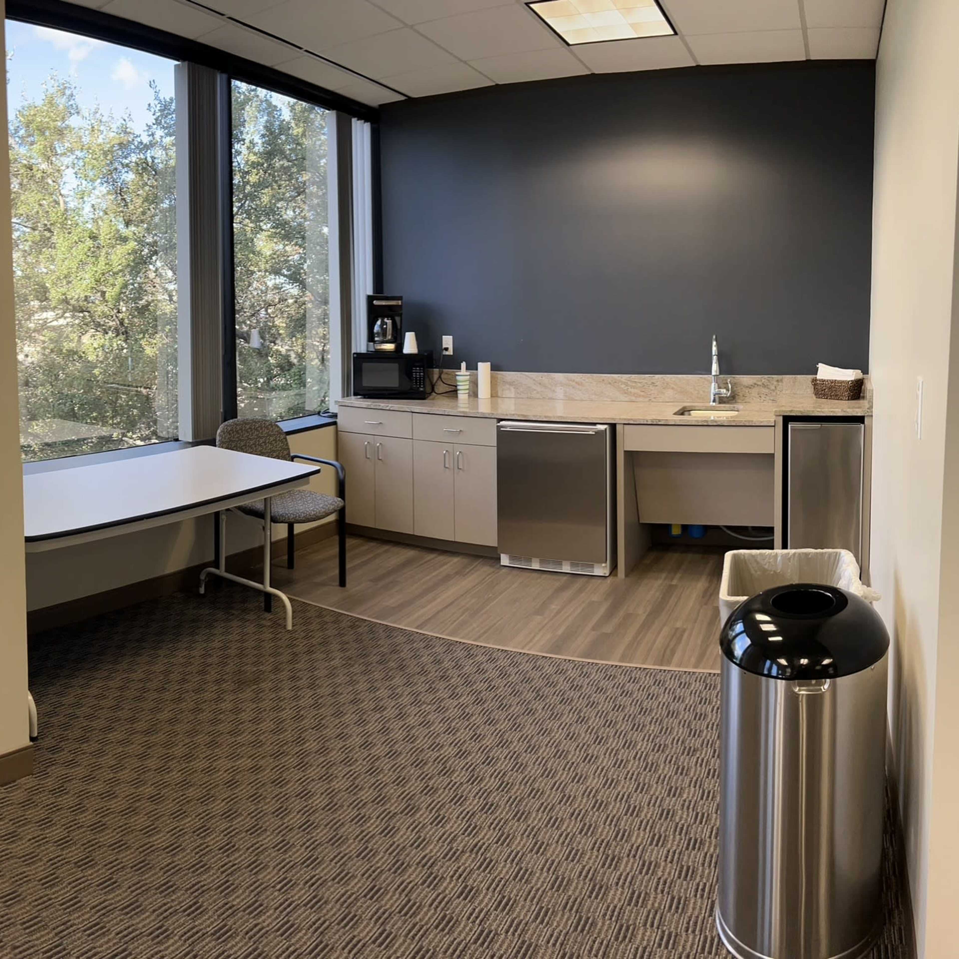 An office break room with a kitchenette, a round table, two chairs, and a large window overlooking greenery outside.