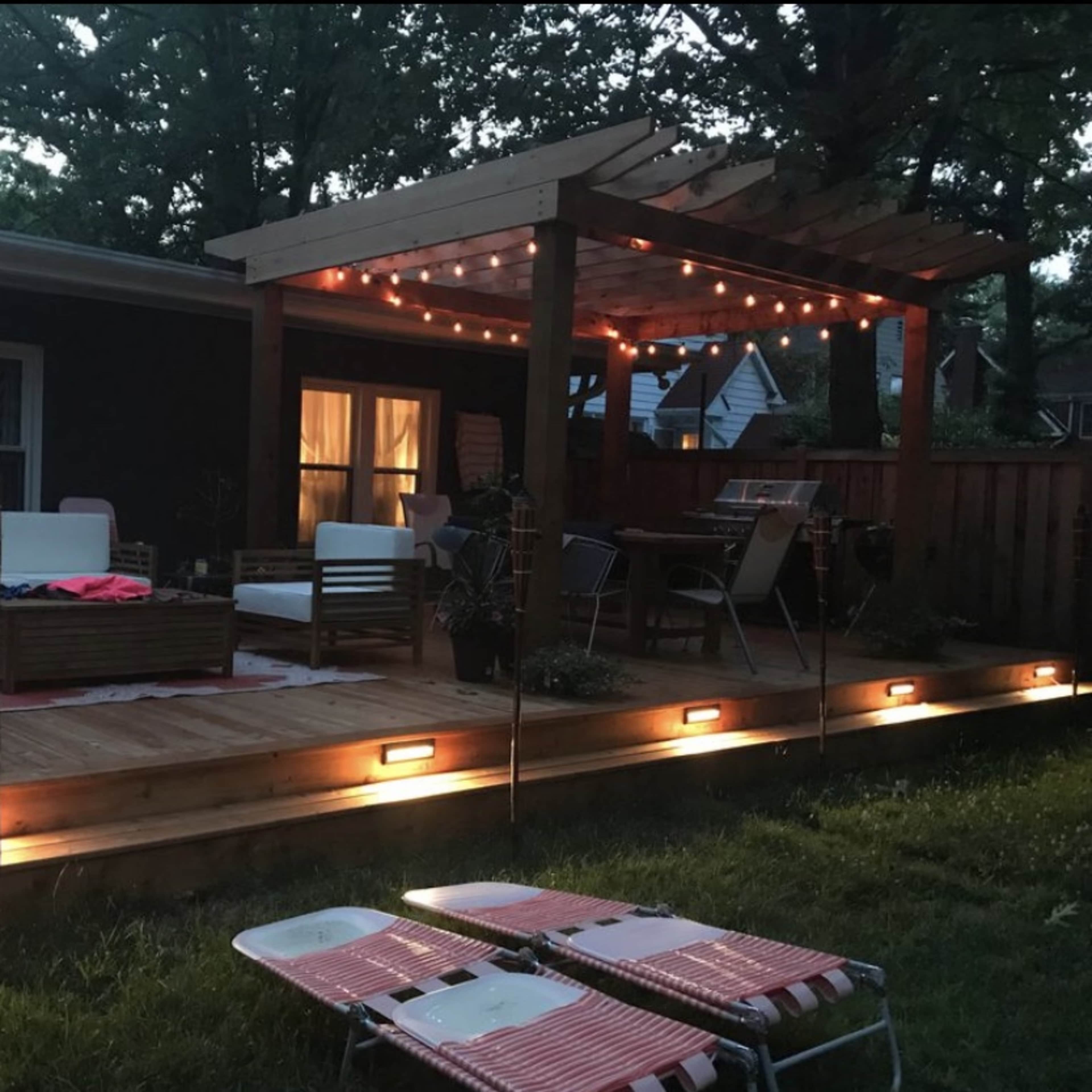 A well-lit wooden deck features a seating area with sofas and a dining table beneath a pergola, surrounded by greenery.