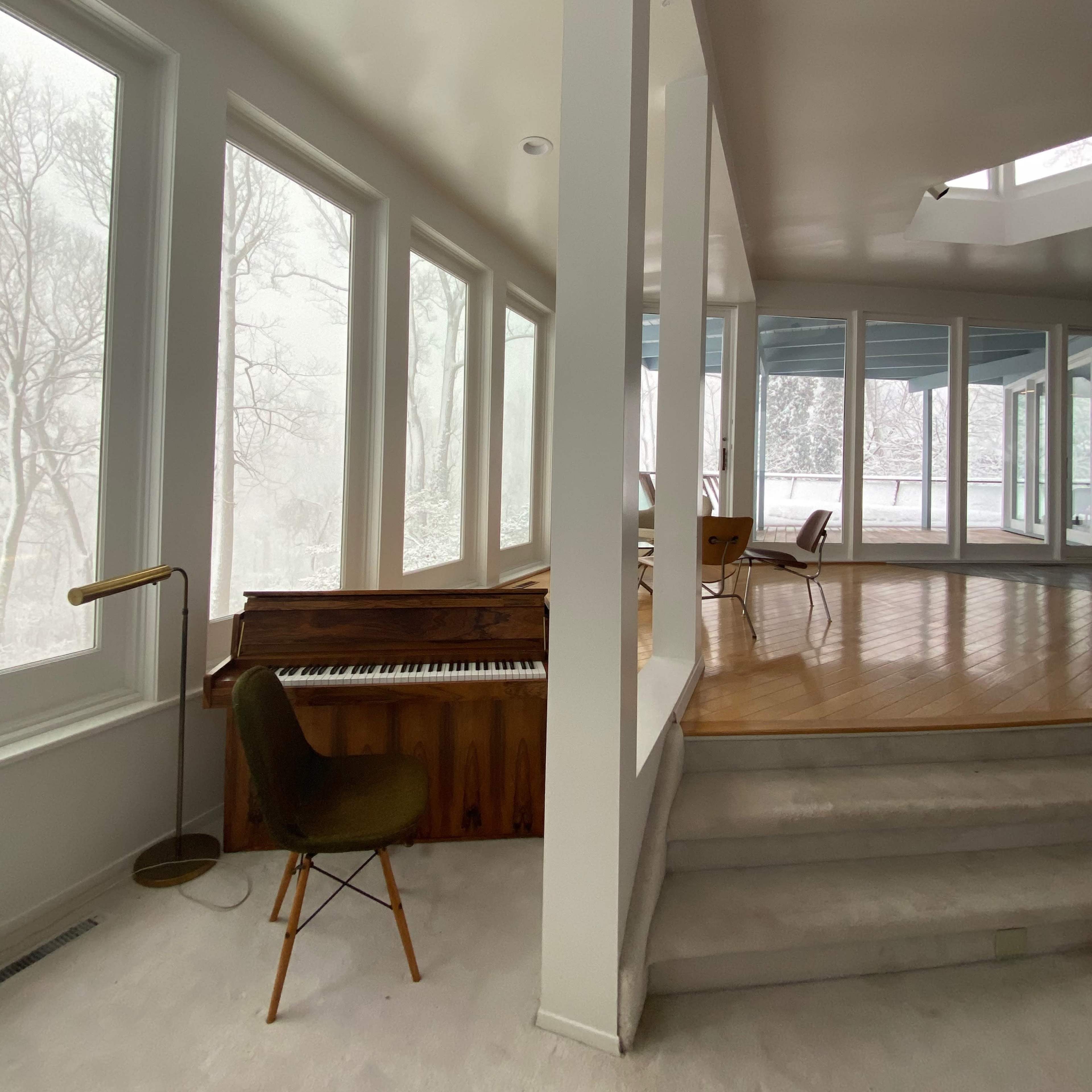 A grand piano sits in a brightly lit room with large windows and a wooden floor.