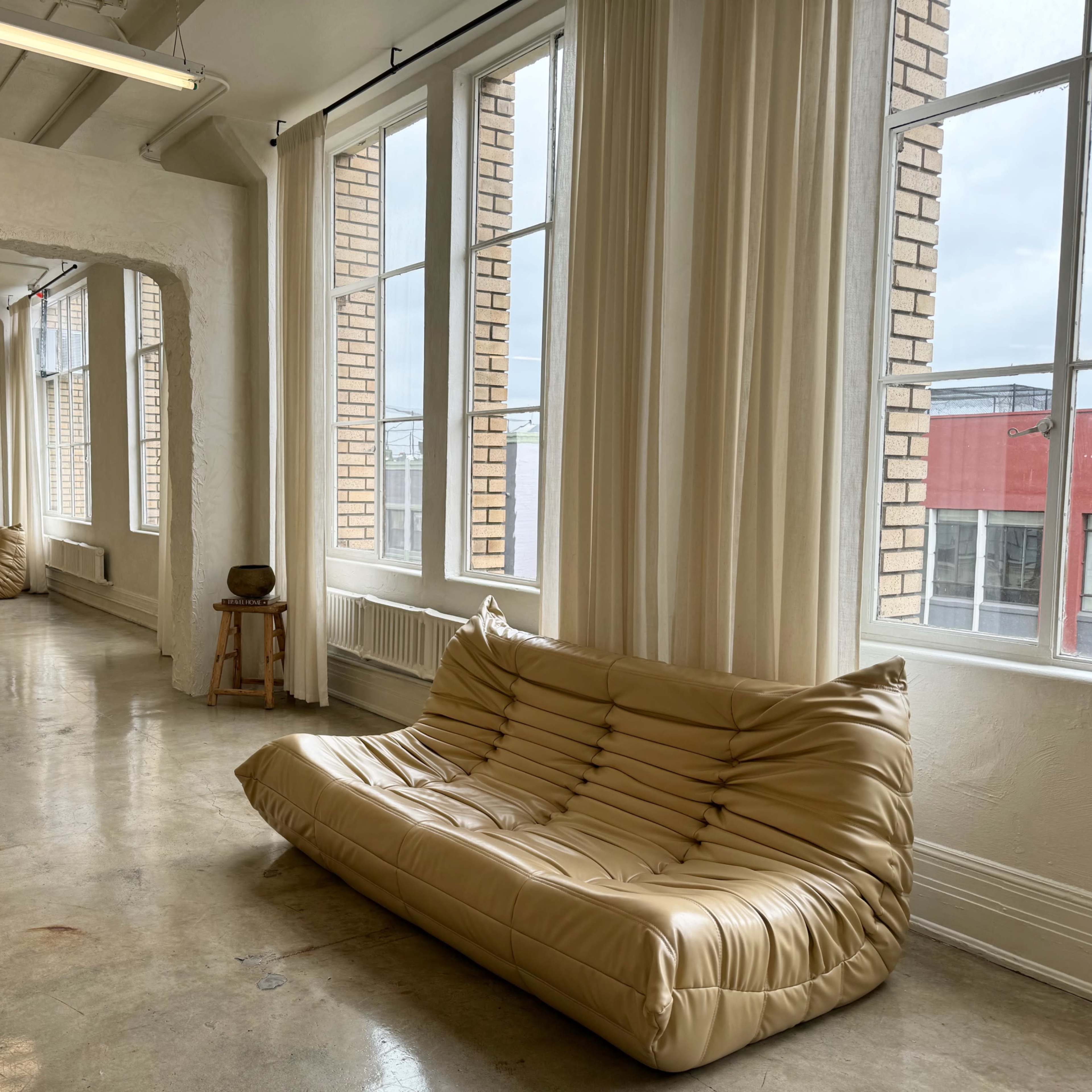 A spacious, brightly lit room featuring large windows, a light-colored sofa, and exposed brick walls.