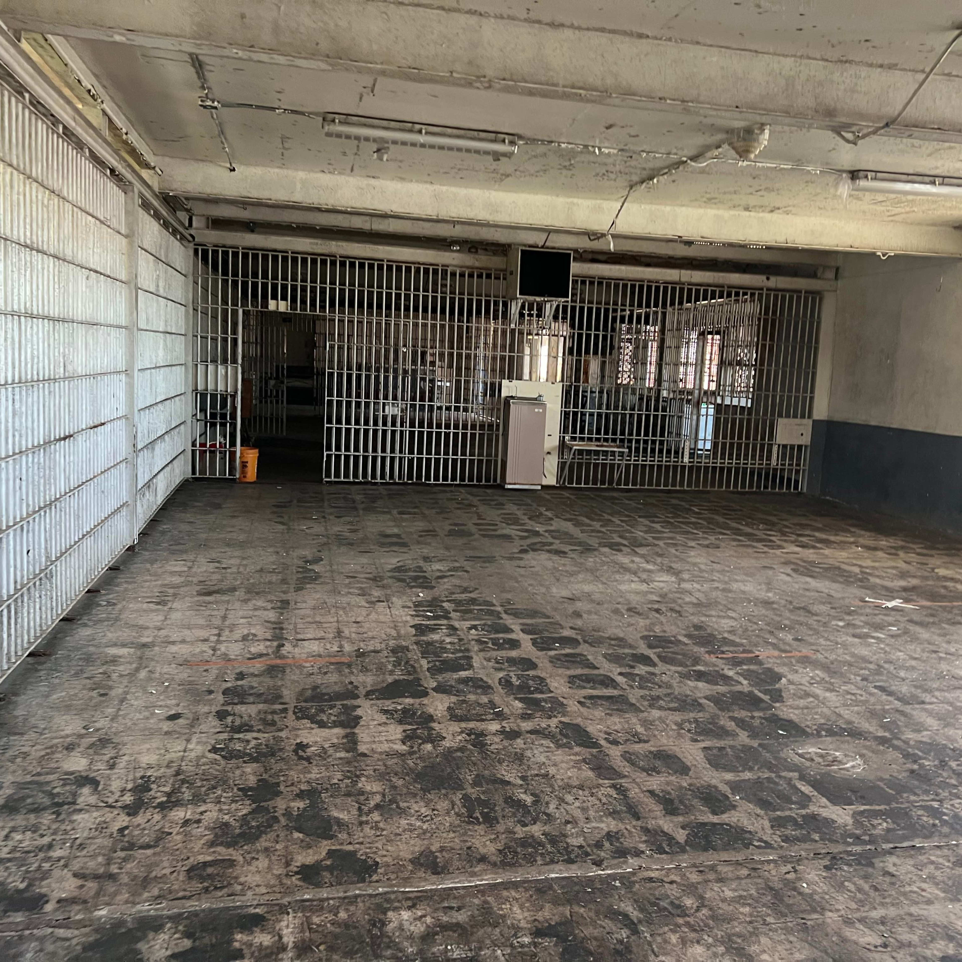 A large, empty room with concrete flooring and barred windows, featuring visible signs of wear on the walls and floor.