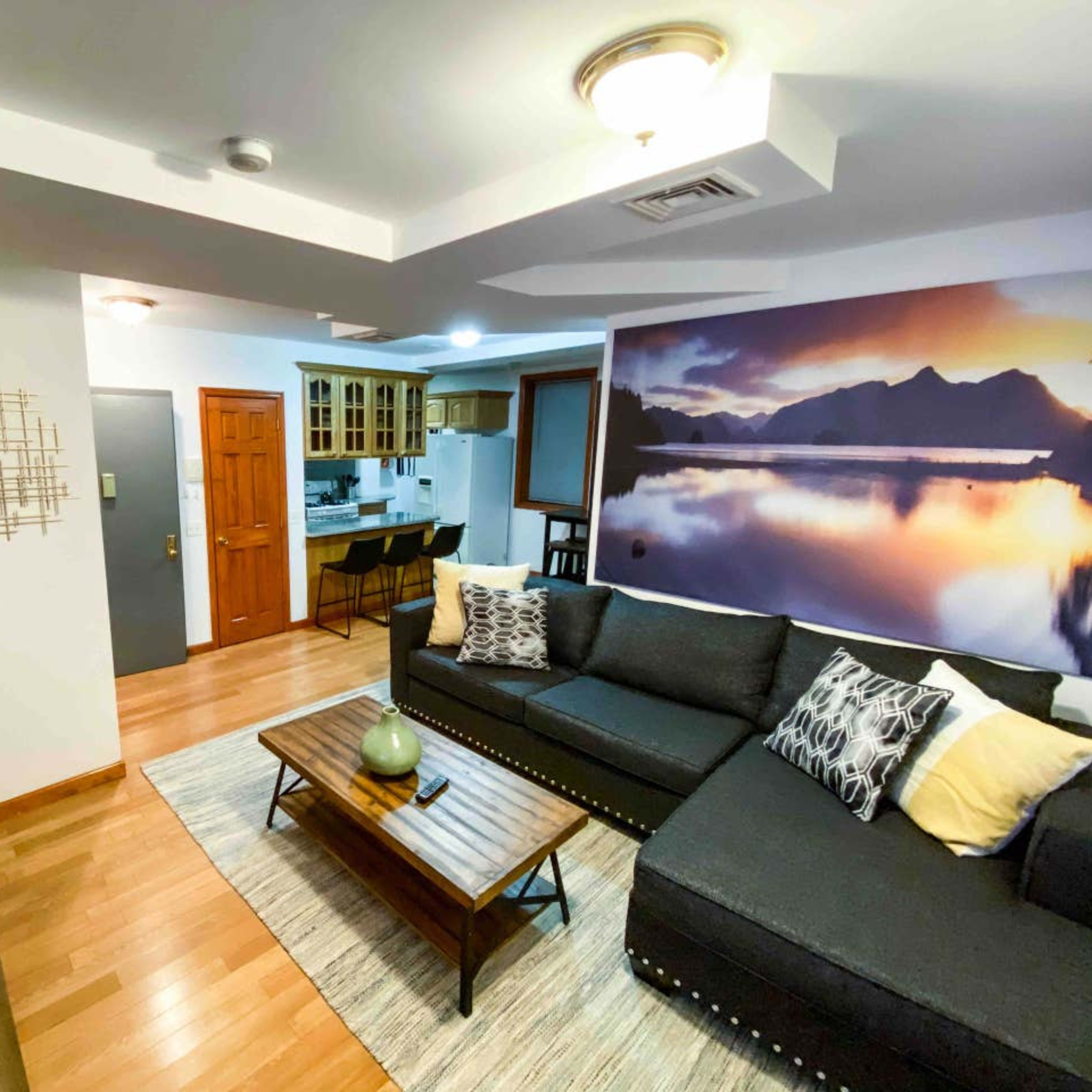 A modern living room features a black sectional sofa, wooden coffee table, and a large wall mural of a mountain landscape, with a dining area visible in the background.