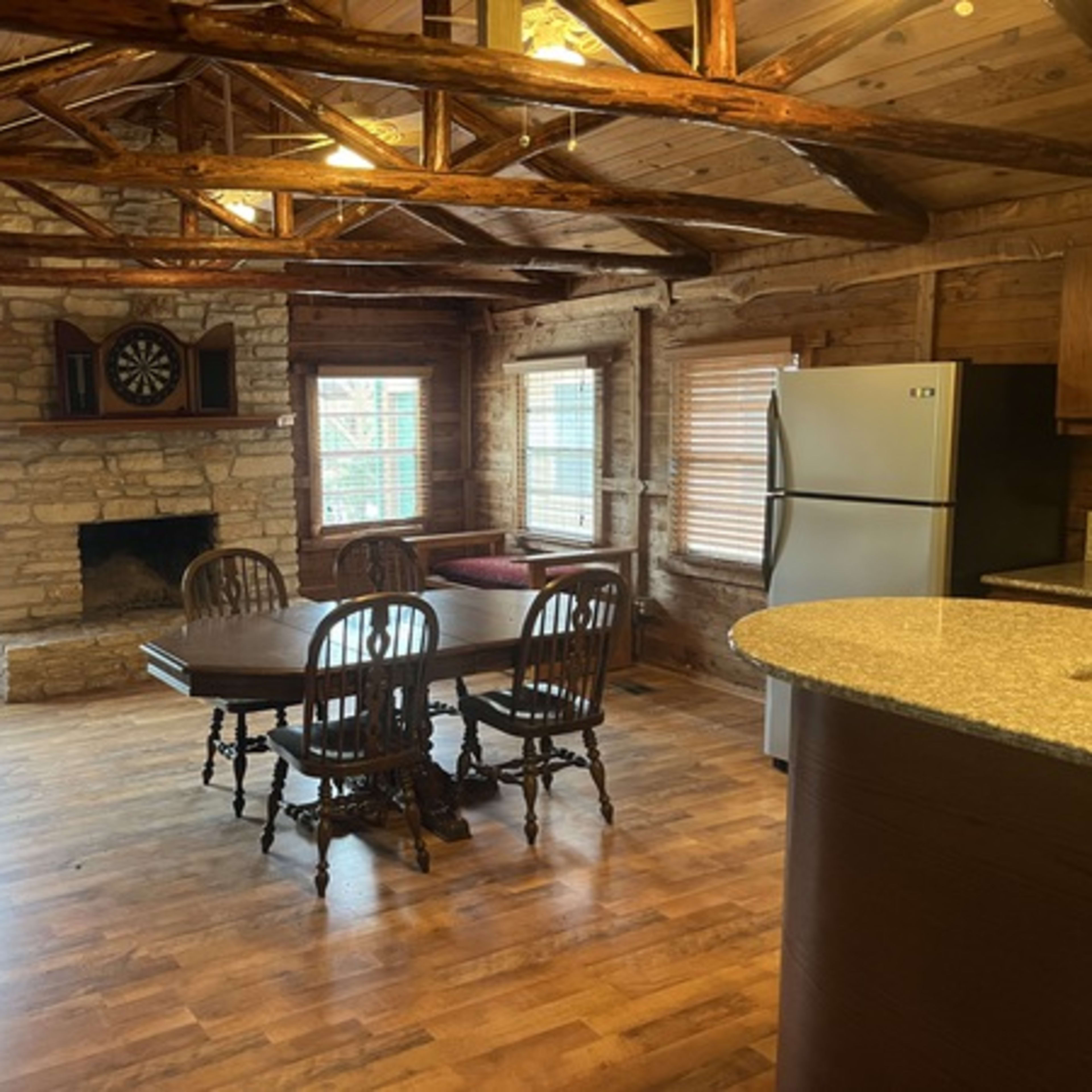 A rustic cabin interior featuring a dining table set for four, a stone fireplace, a refrigerator, and wooden beams on the ceiling.