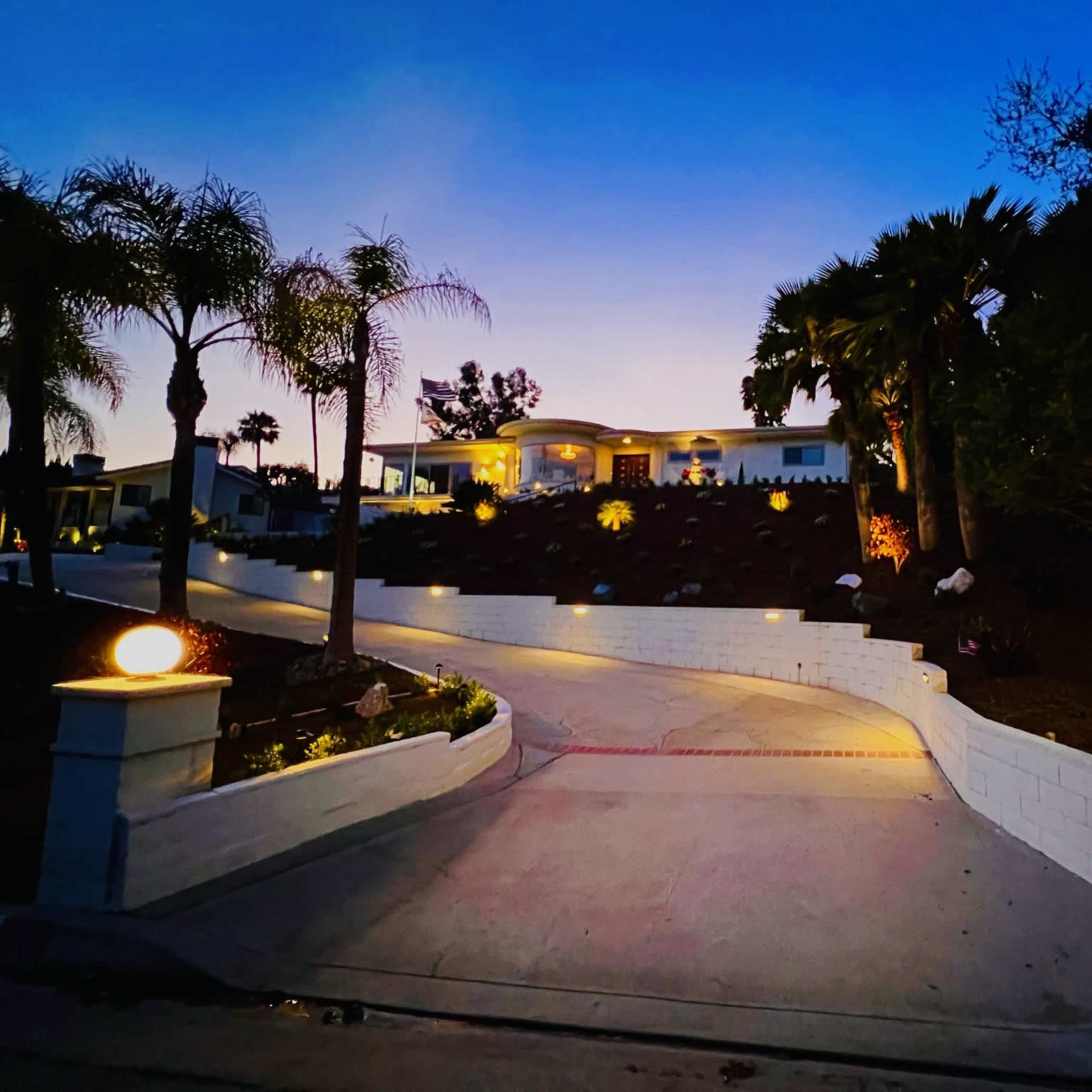 A well-lit driveway leads up to a modern house surrounded by palm trees and landscaping against a twilight sky.