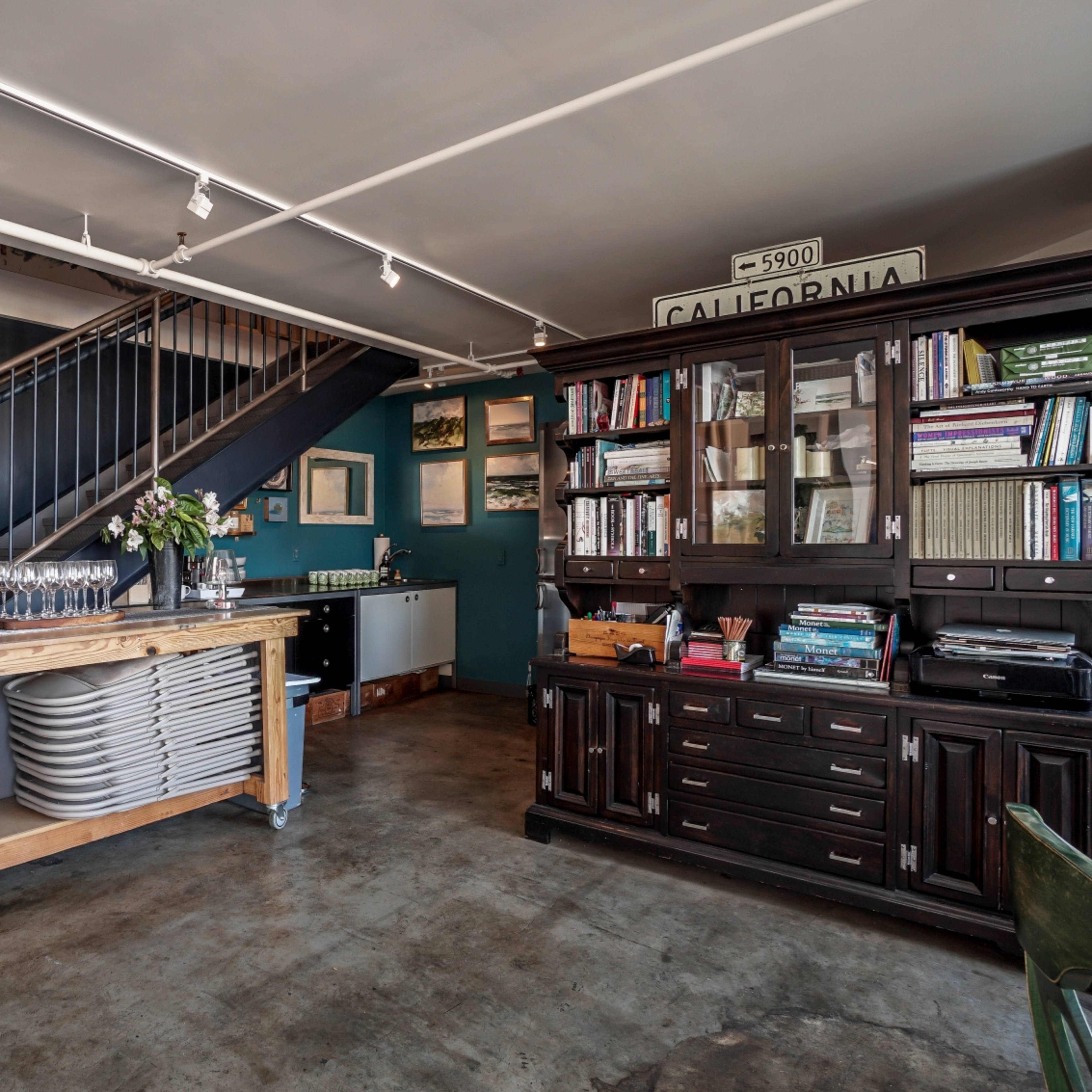 The interior of a cafe features a wooden table with stacked chairs, a dark wooden bookshelf filled with books, and a staircase in the background.