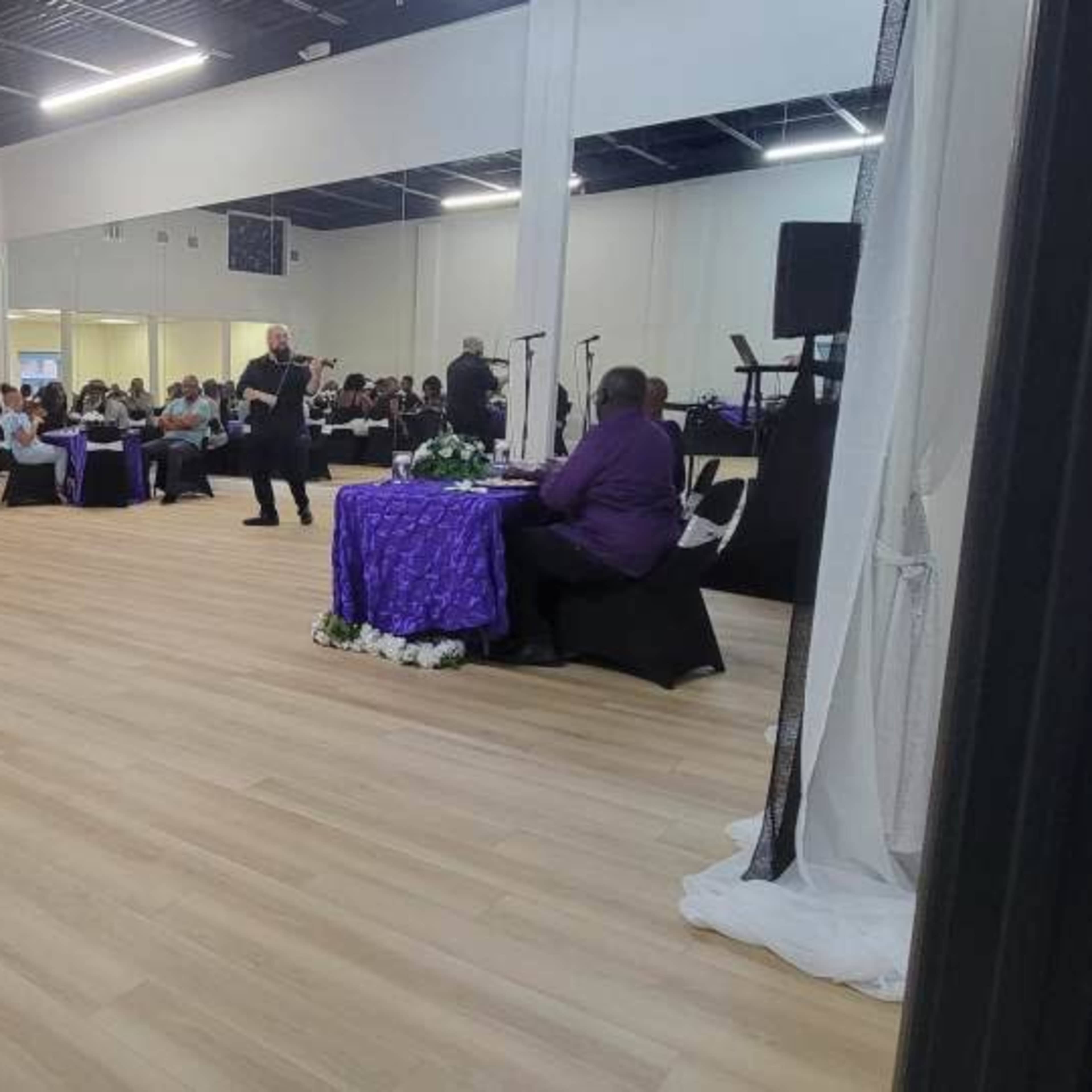 A large indoor space set up for an event with tables arranged in a dining format, featuring guests seated at various tables adorned with white and purple tablecloths.