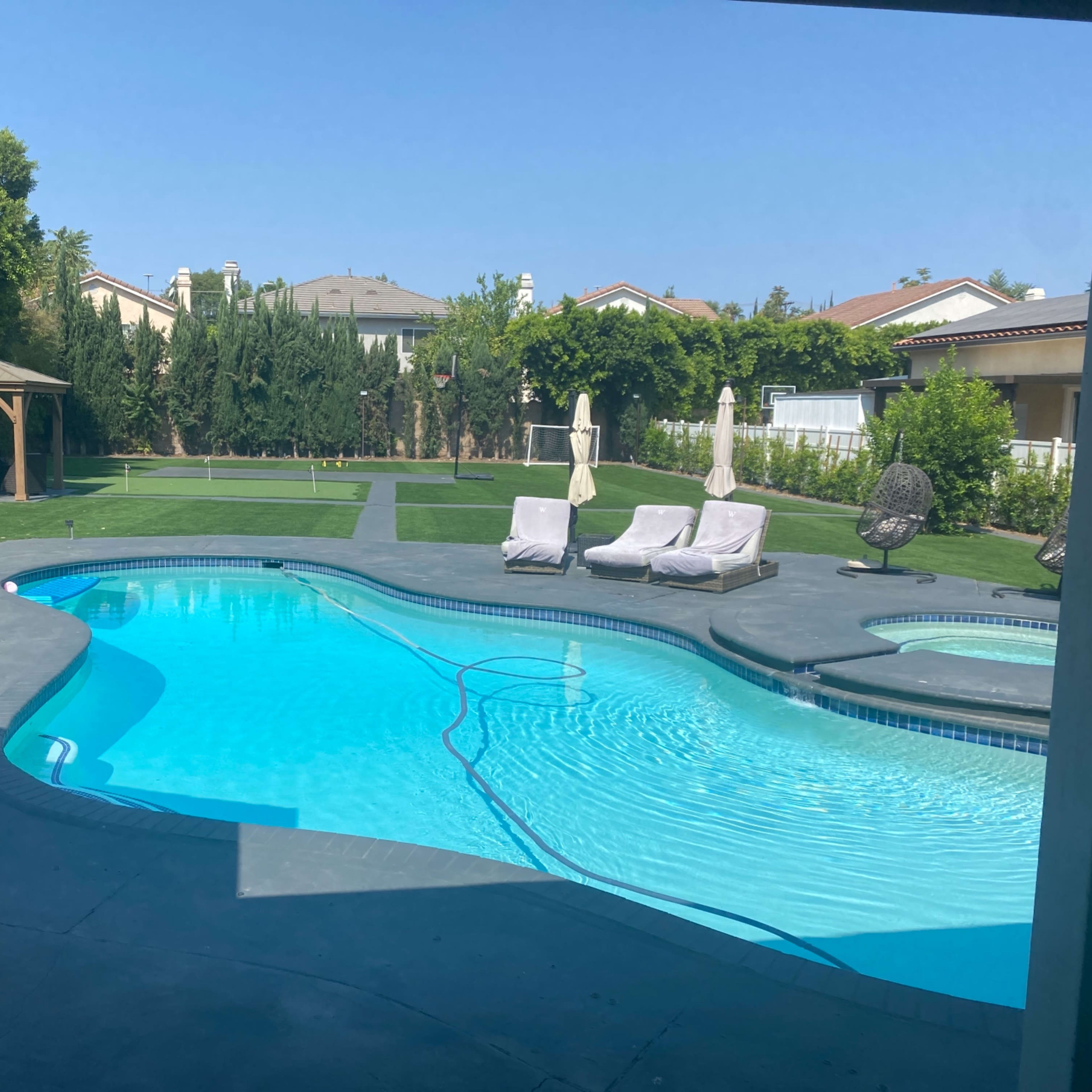 A backyard with a swimming pool surrounded by lounge chairs and grass areas, featuring a gazebo in the background.