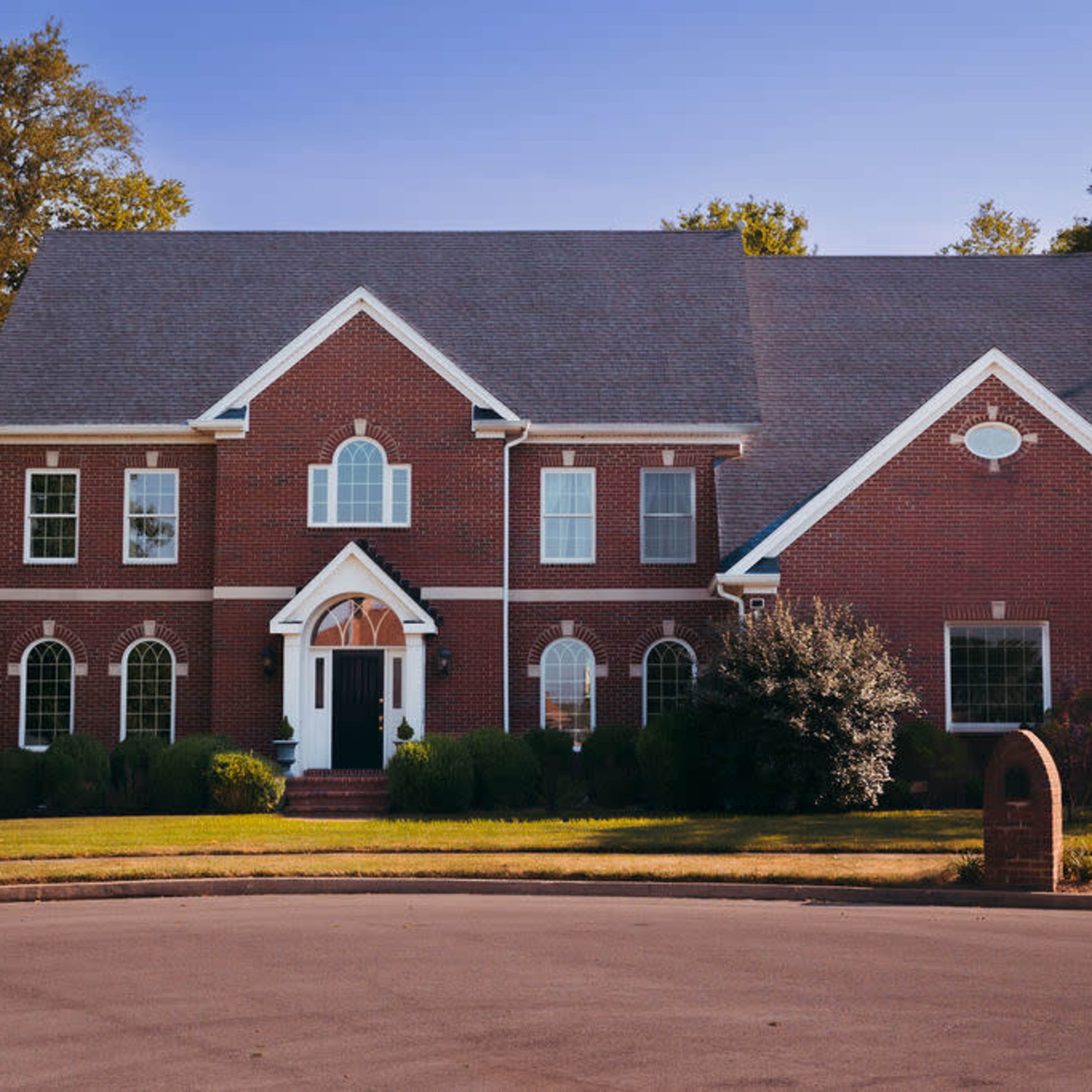 A two-story brick house with a gabled roof and multiple windows is situated on a corner lot with a landscaped front yard.