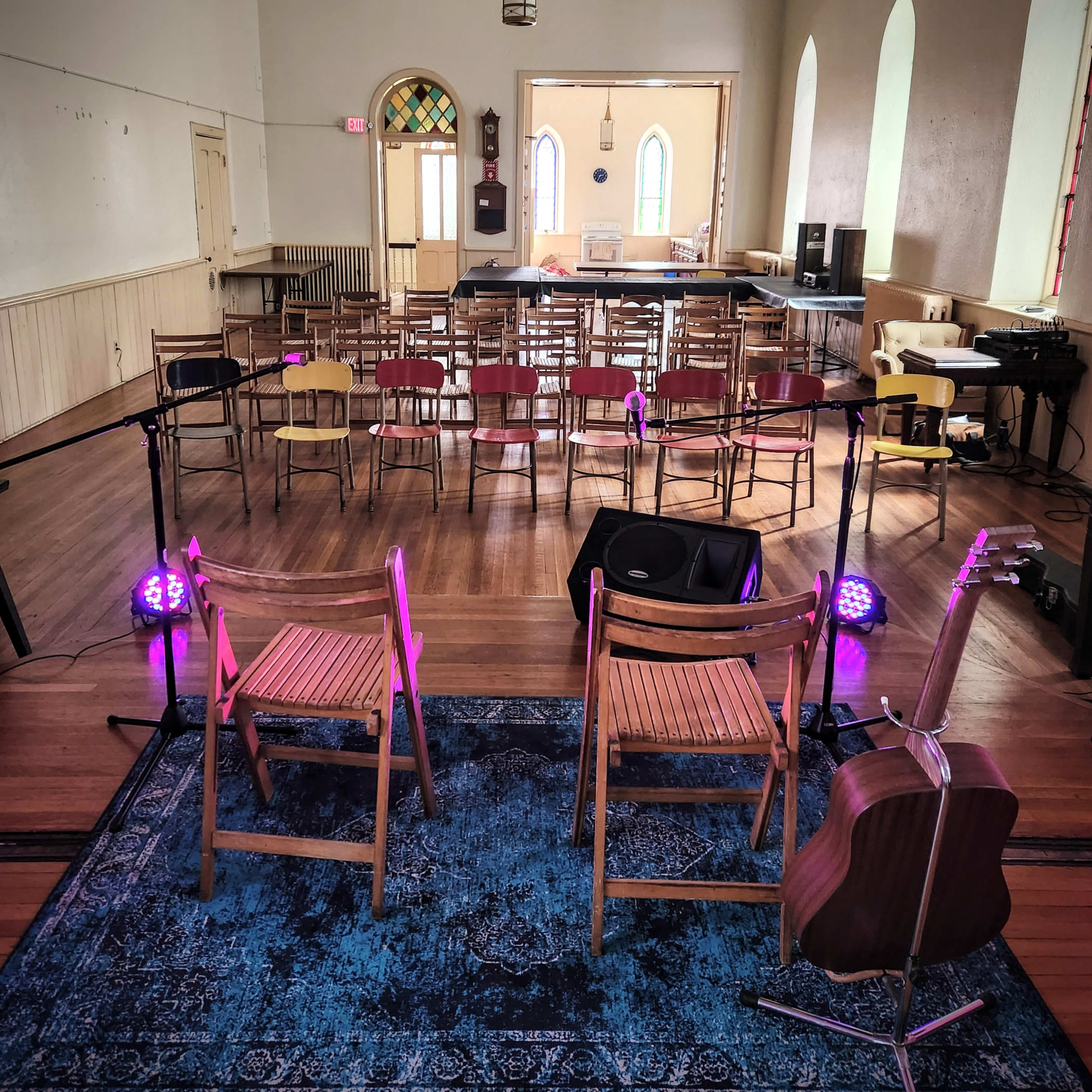 A small performance space with two wooden chairs facing an array of chairs set up for an audience, accompanied by a guitar on a stand and stage lighting.