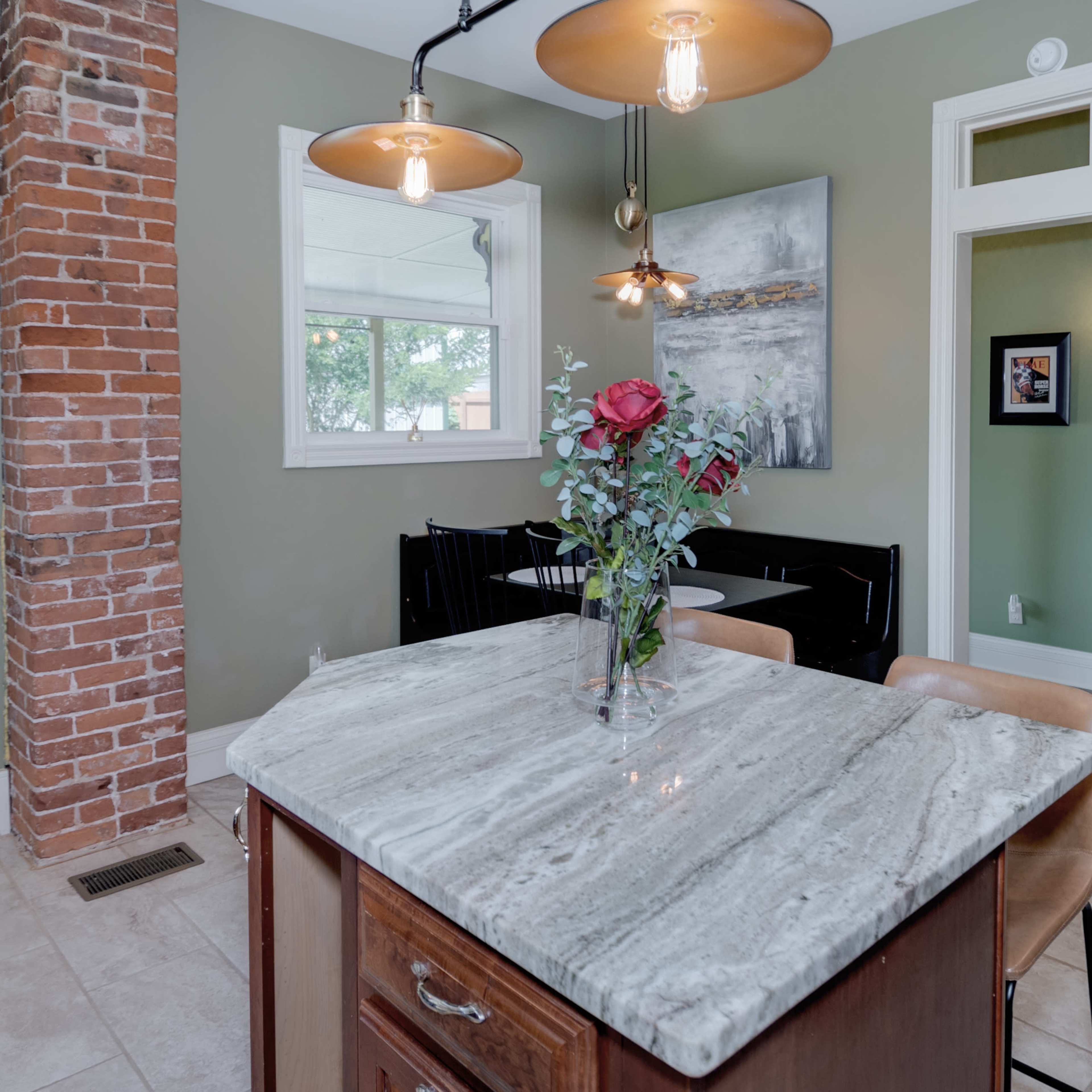 A kitchen features a marble island with a vase of roses, surrounded by a dining area and a brick pillar.