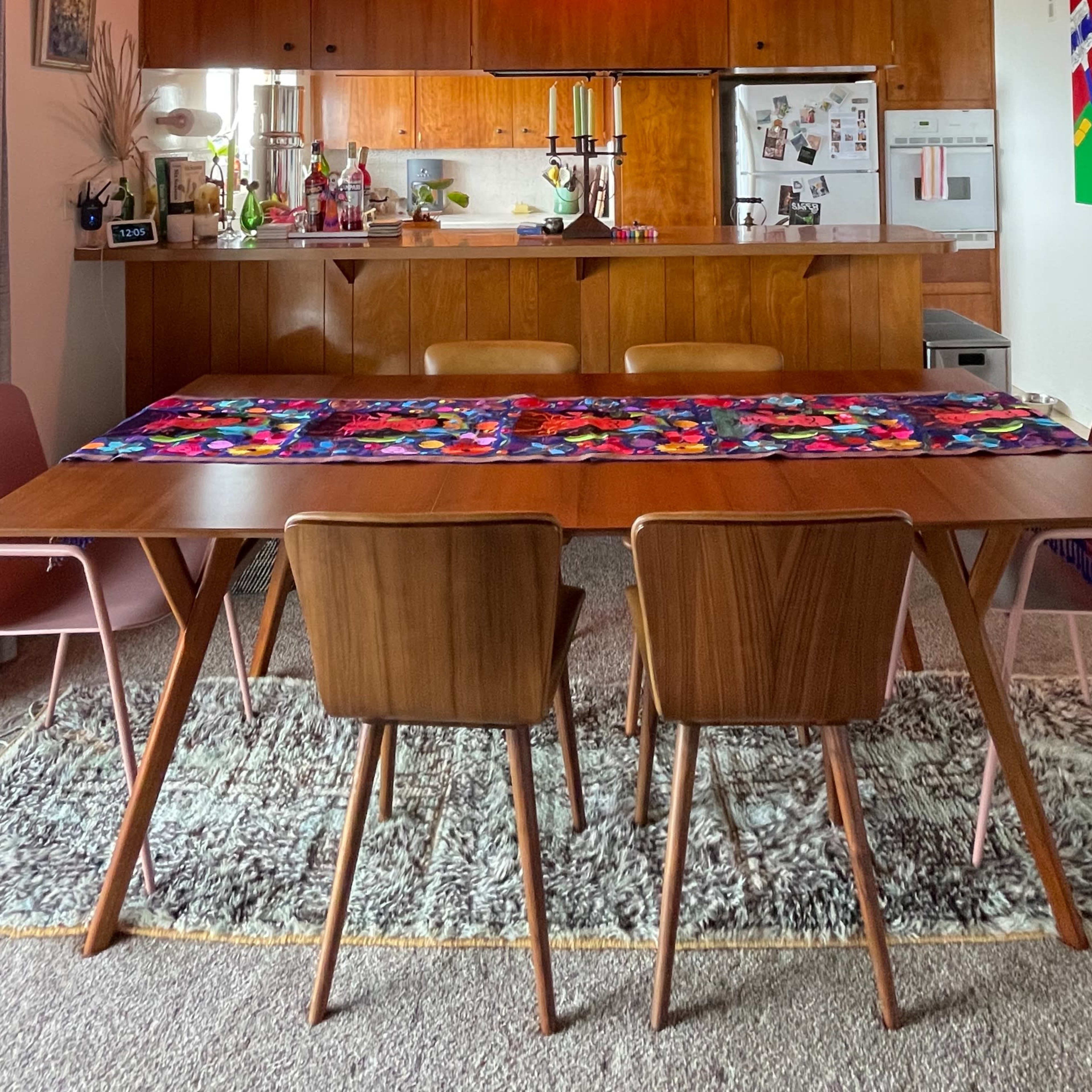 A dining table with four chairs is set in a brightly lit room featuring a kitchen area in the background.