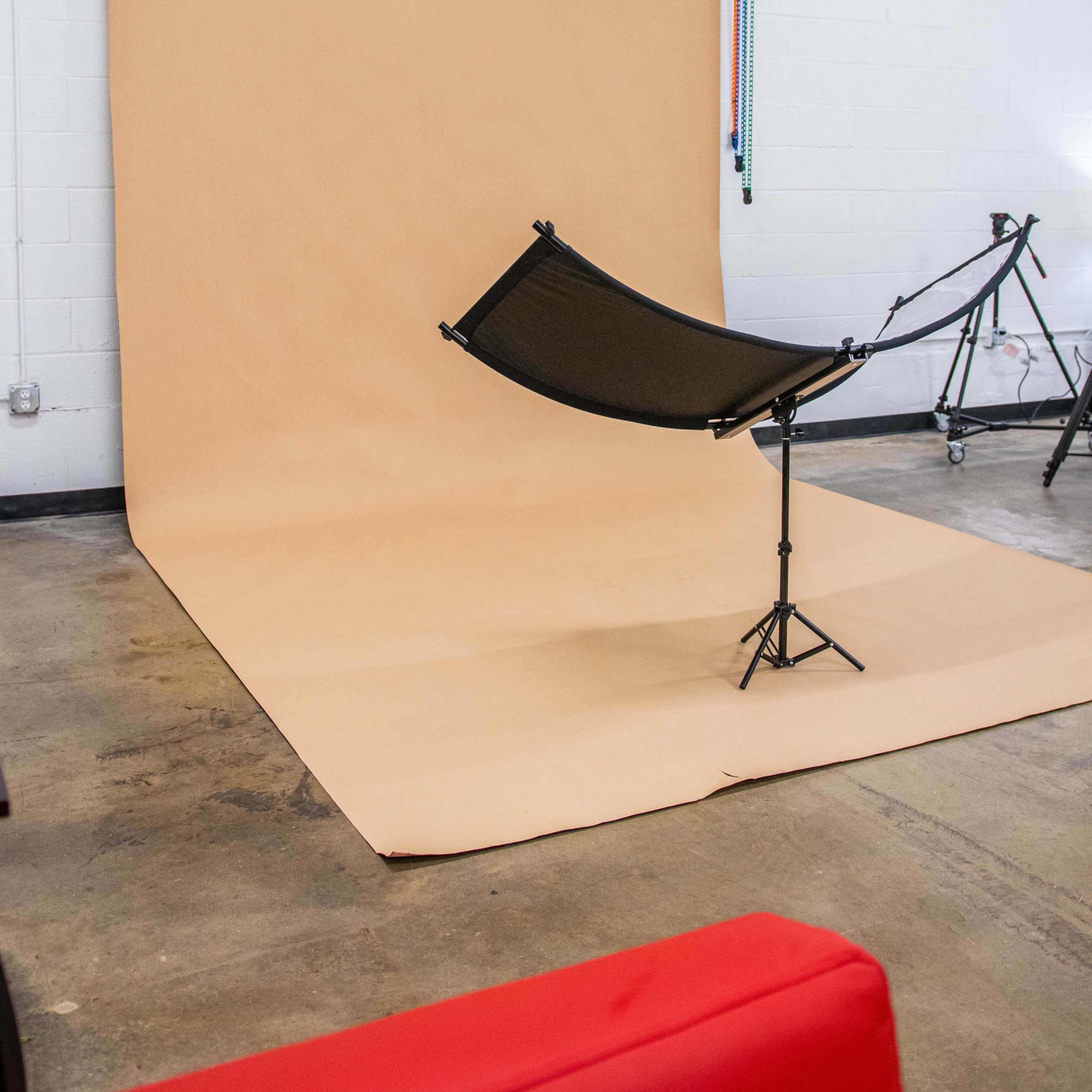 A photography studio setup features a tan backdrop with colored rolls above and a black fabric hammock suspended on a stand in the foreground.
