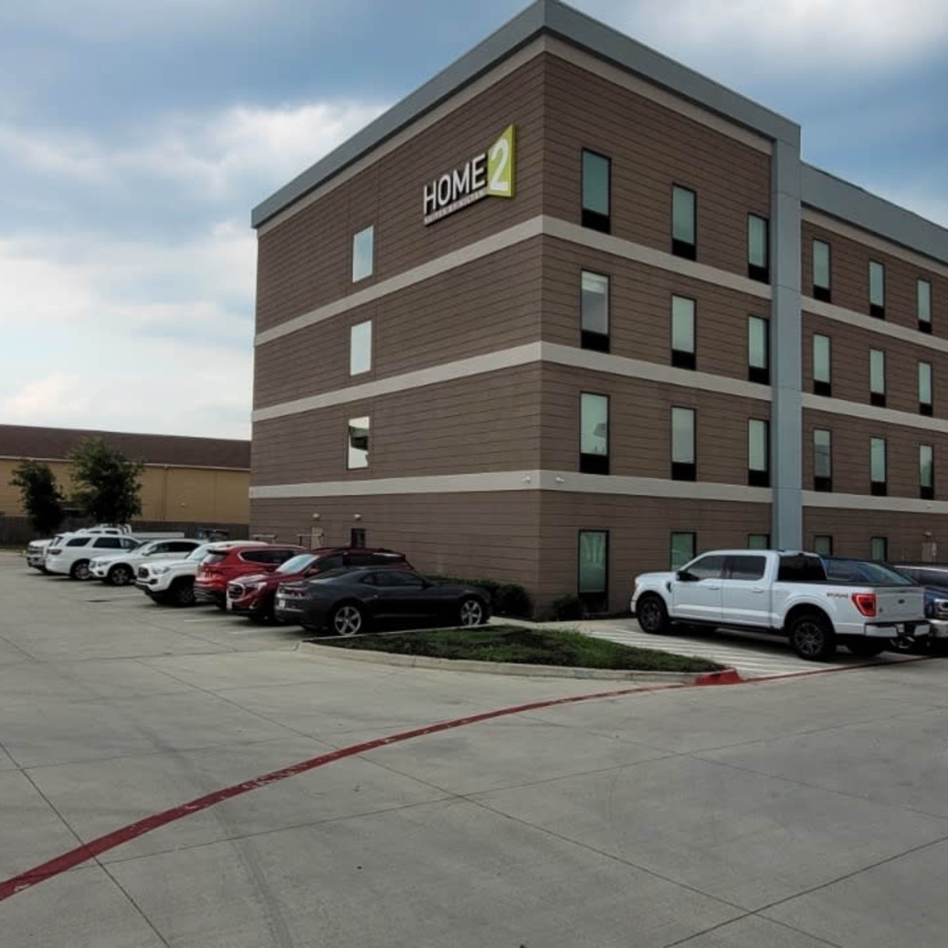 A multistory hotel building next to a parking lot with several vehicles, set against a cloudy sky.