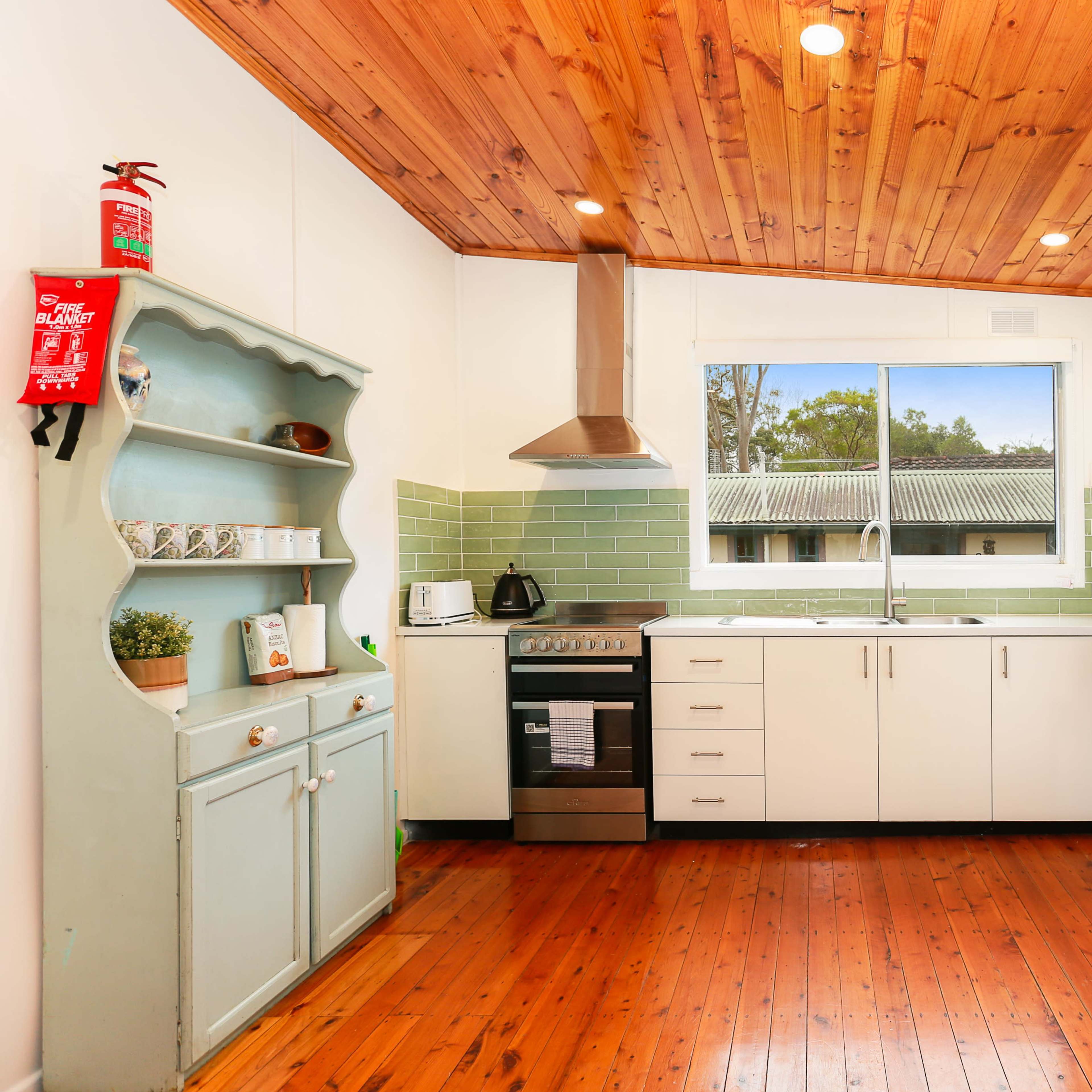 The kitchen features wooden flooring, white cabinetry, a stainless steel stove, and a light green tiled backsplash next to a window.