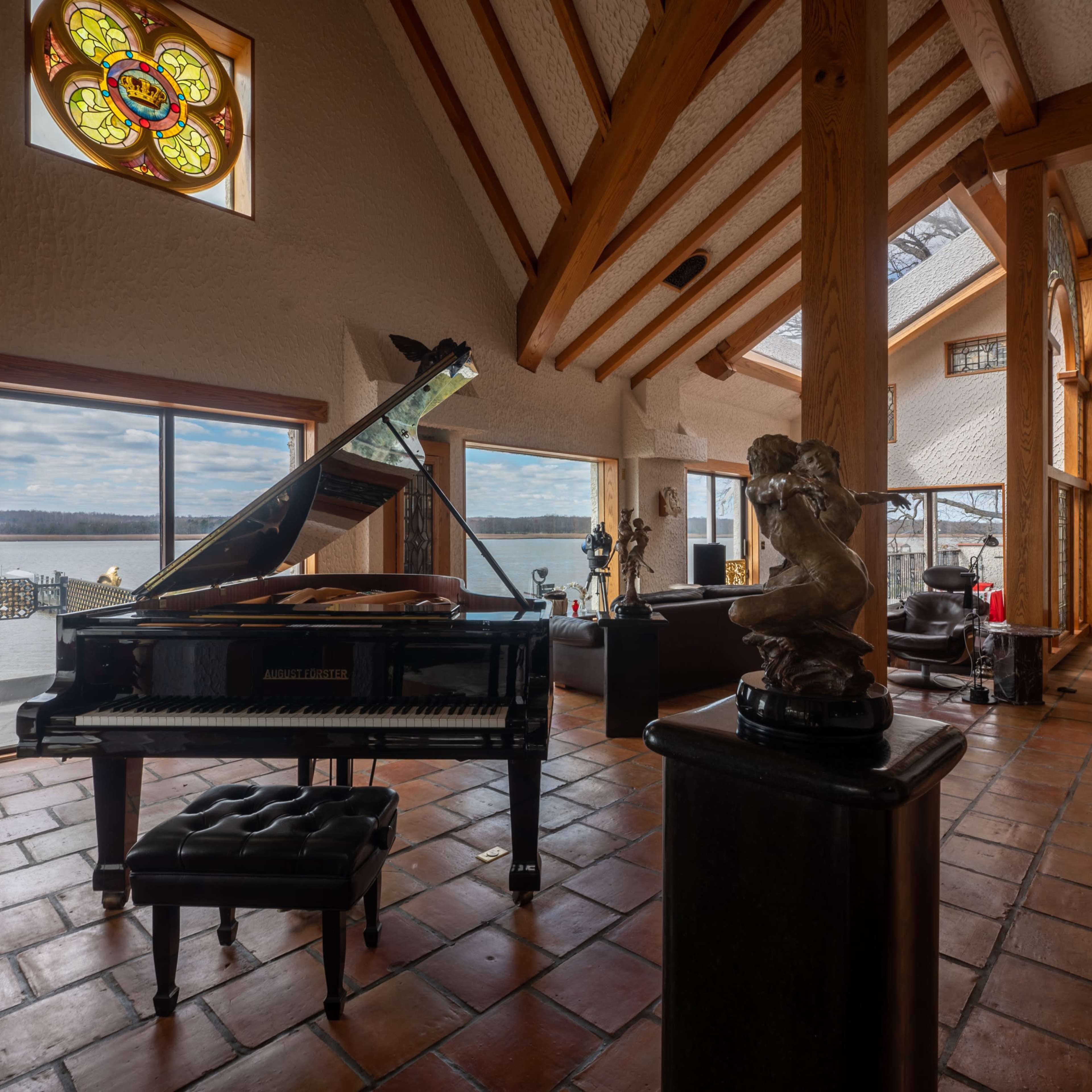 A grand piano sits in a sunlit room with large windows overlooking a lake, surrounded by wooden beams and sculptures.