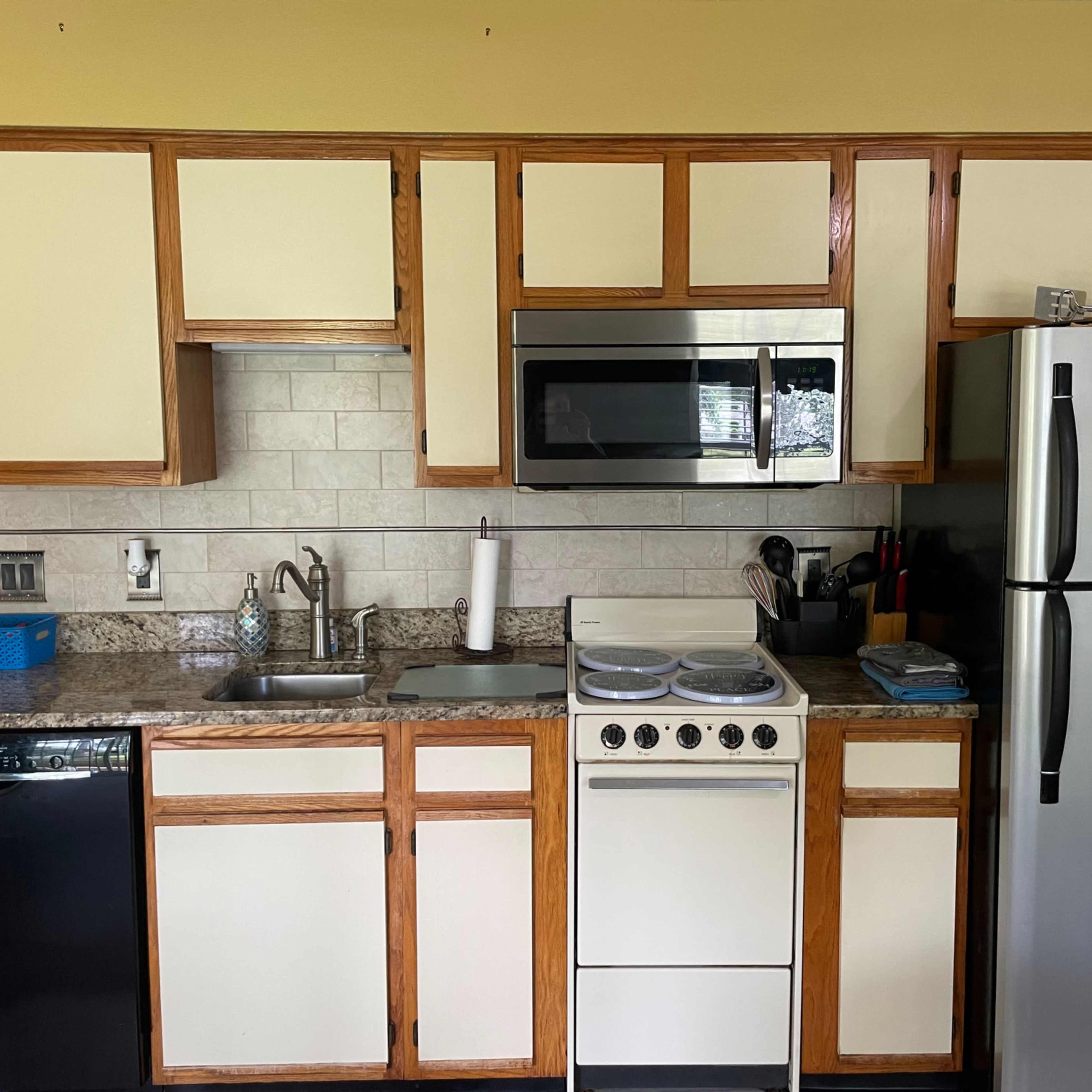 A kitchen with wooden cabinets, a stainless steel refrigerator, a stove, a microwave, and a sink with various utensils and appliances on the counter.