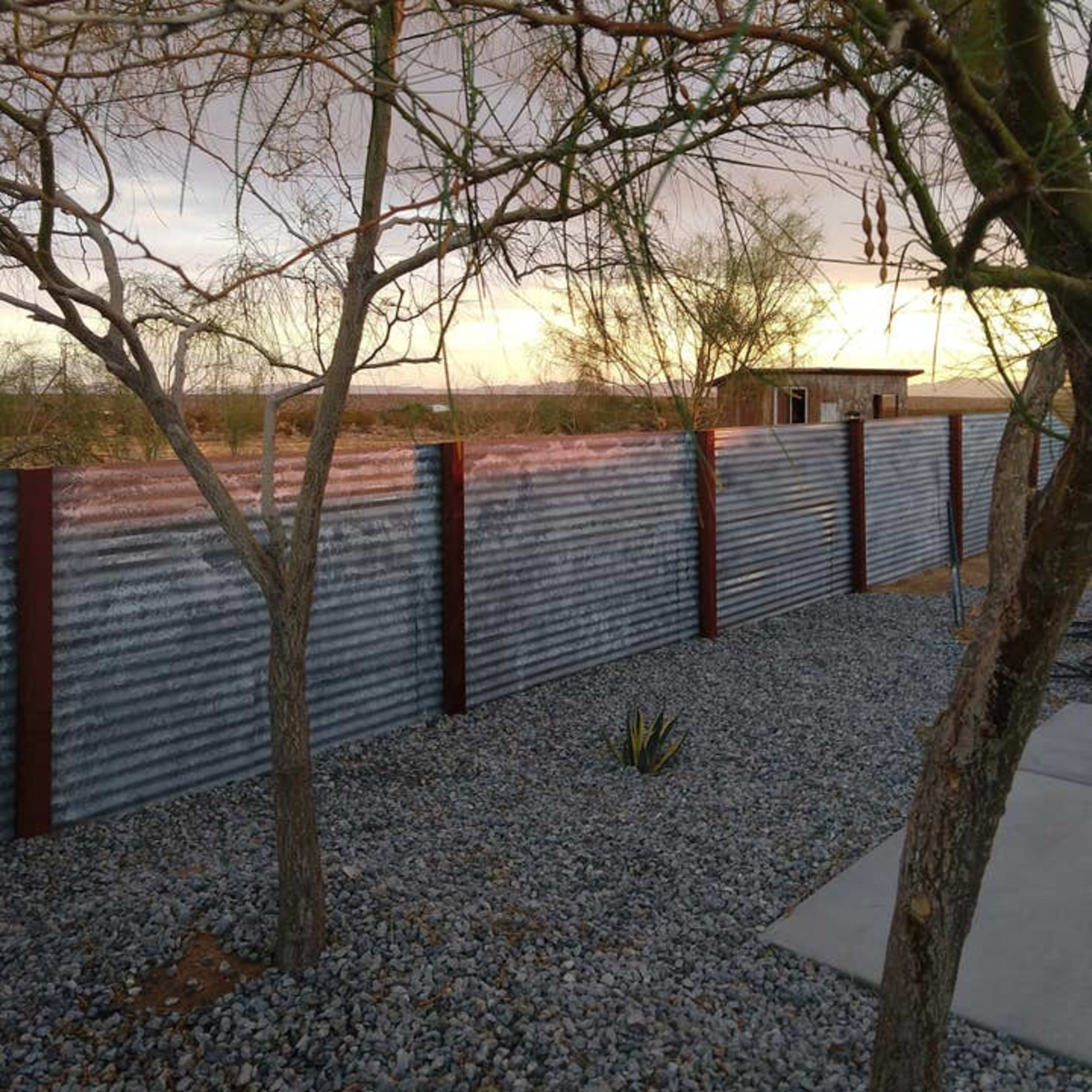 A corrugated metal fence surrounded by gravel and a few sparse trees against a cloudy sky.