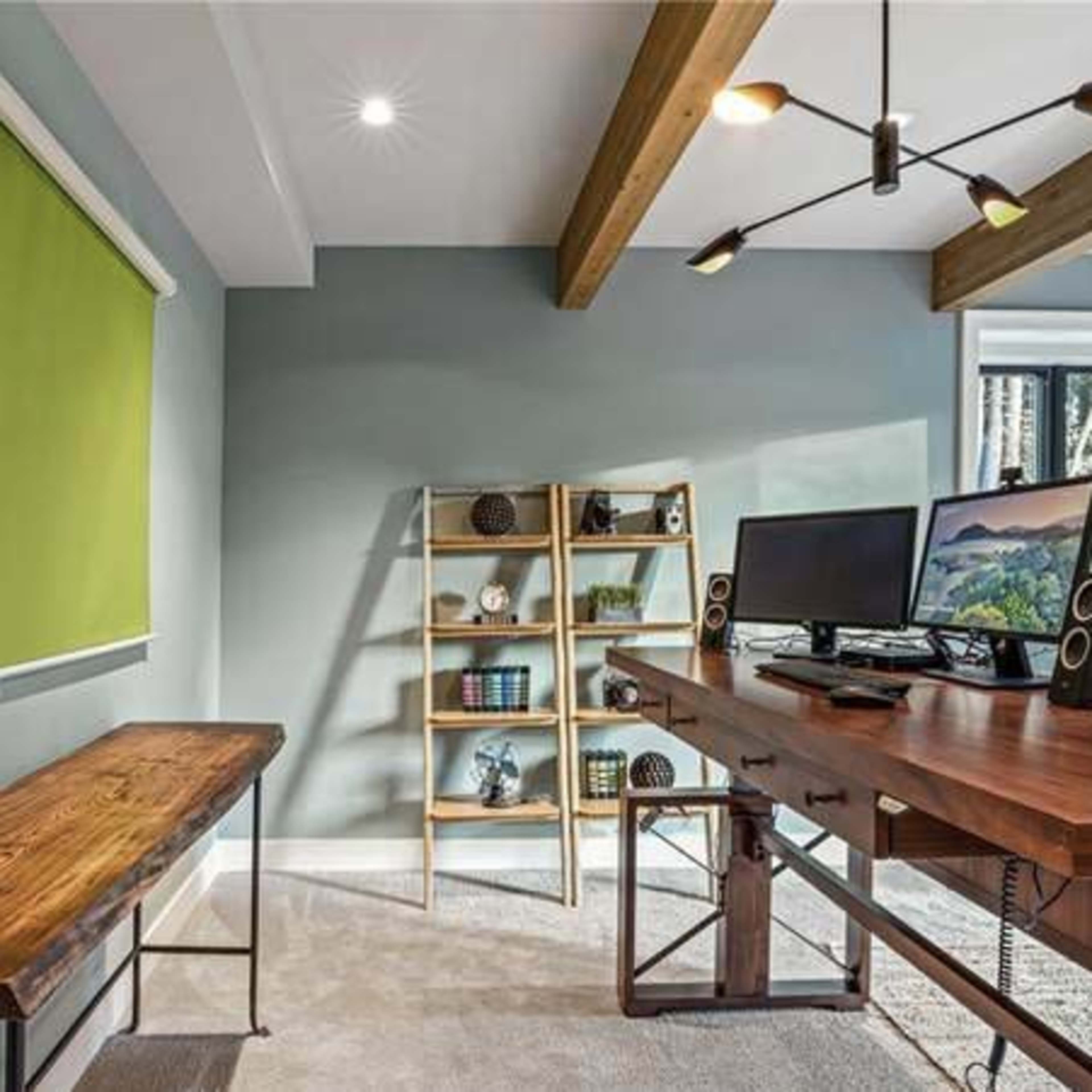 A modern office space featuring a green wall-mounted screen, a wooden bench, a desk with two monitors, and a shelving unit with decorative items.