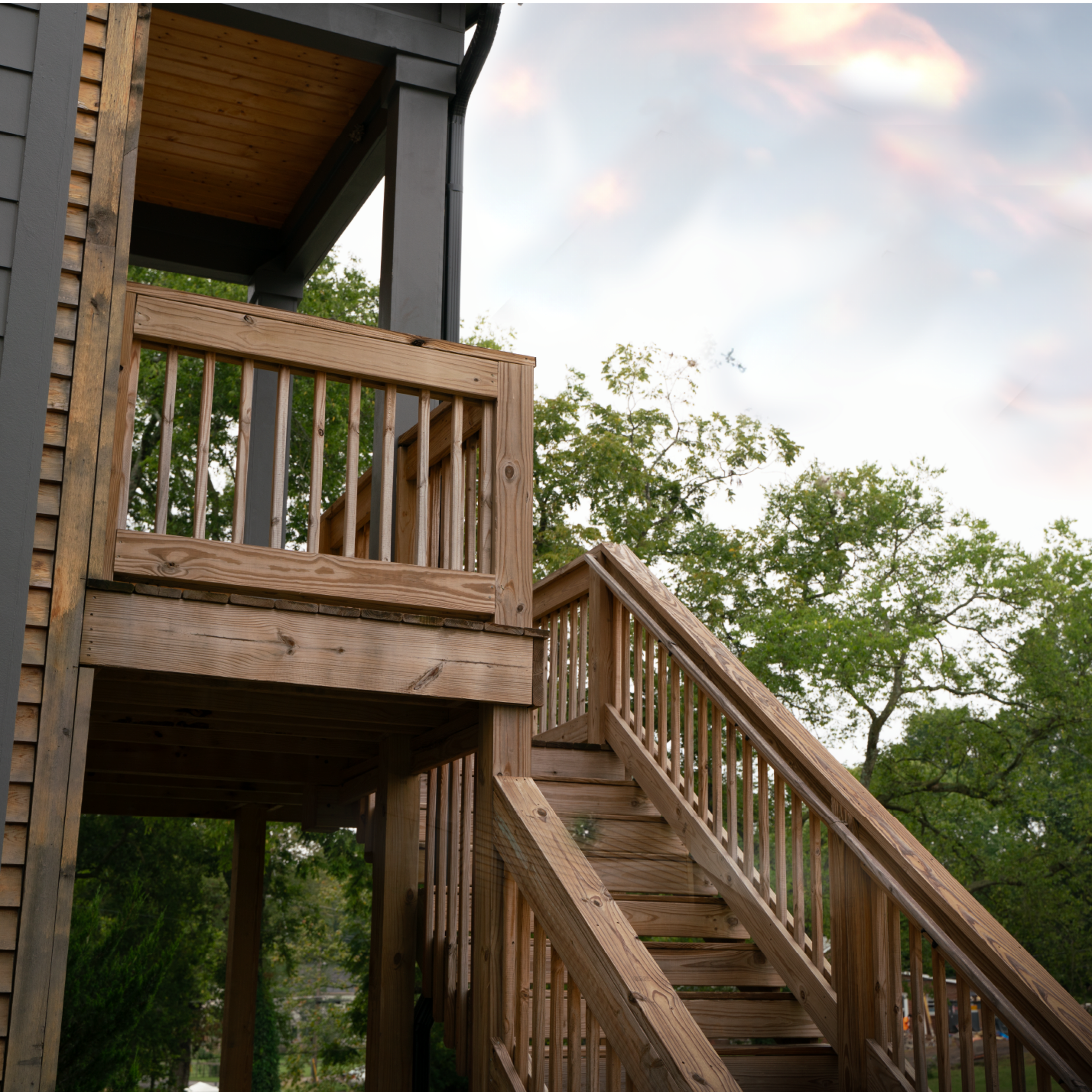A wooden staircase leads to a balcony on the side of a two-story building surrounded by greenery.