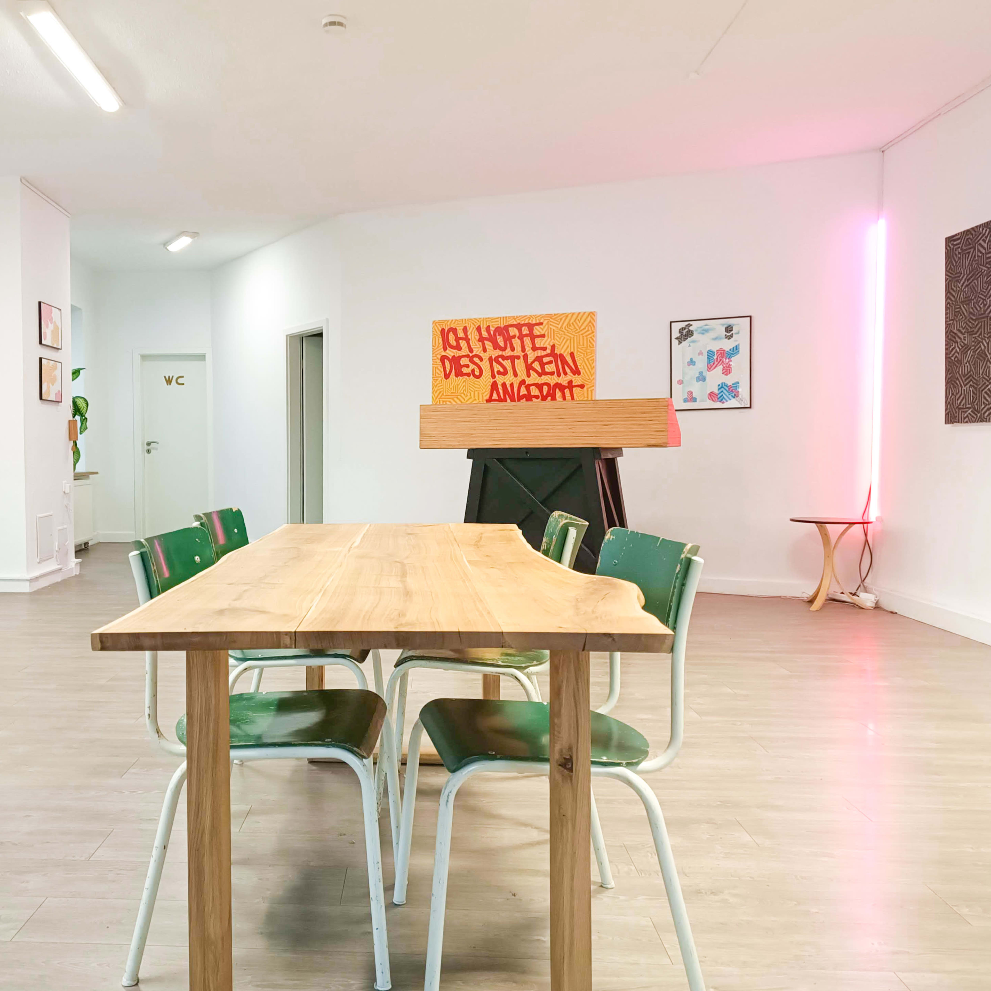 A spacious, modern interior with a wooden table and green chairs, featuring a brightly lit sign that reads "ICH HOFFE, DAS IST KEIN ANSITZ" against a minimalist backdrop.