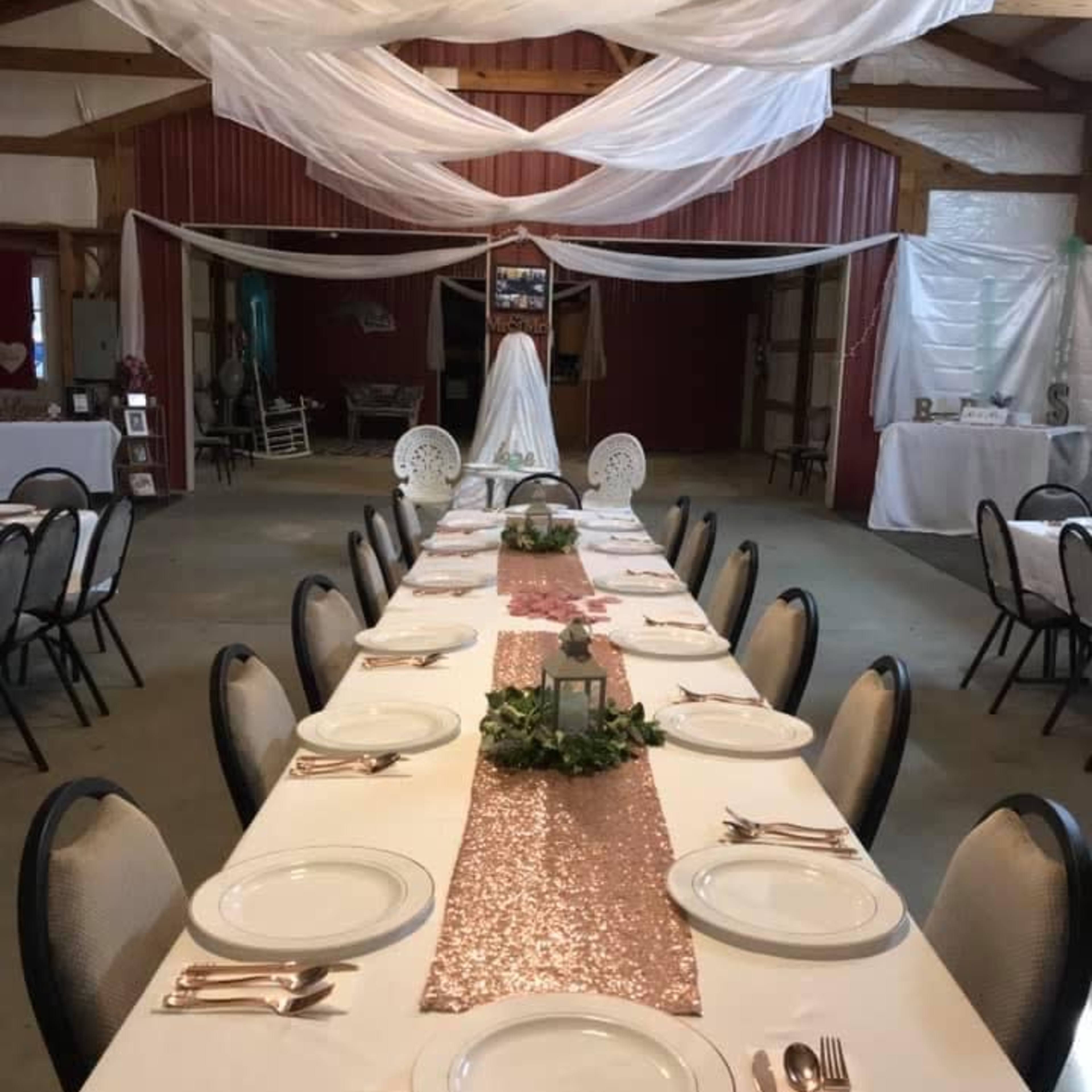 A long banquet table is set with white plates and silverware, adorned with a sparkling rose gold runner and greenery, under draped fabric in a rustic venue.