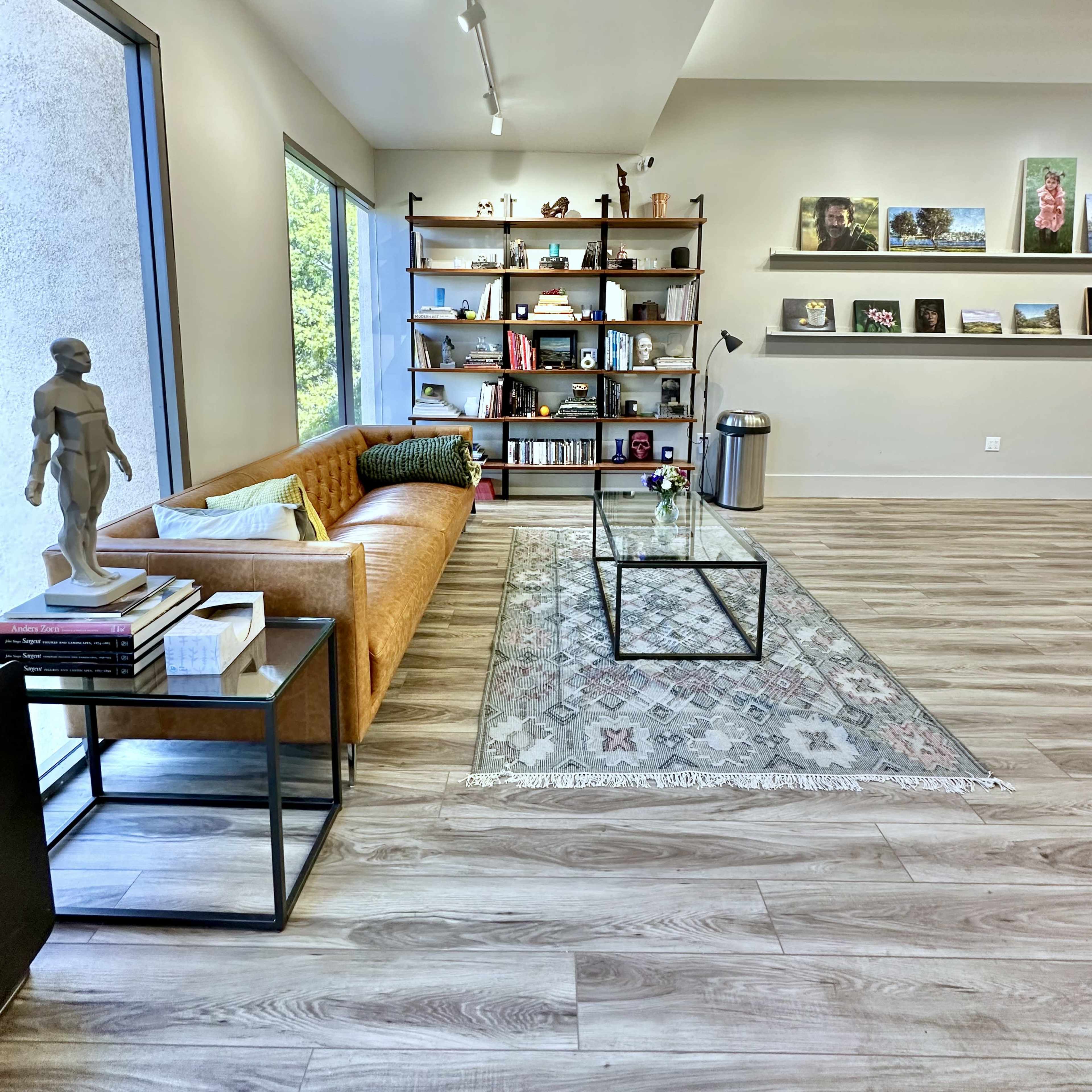 A modern living space with a leather sofa, a glass coffee table, a bookshelf filled with books and decorative items, and large windows allowing natural light.