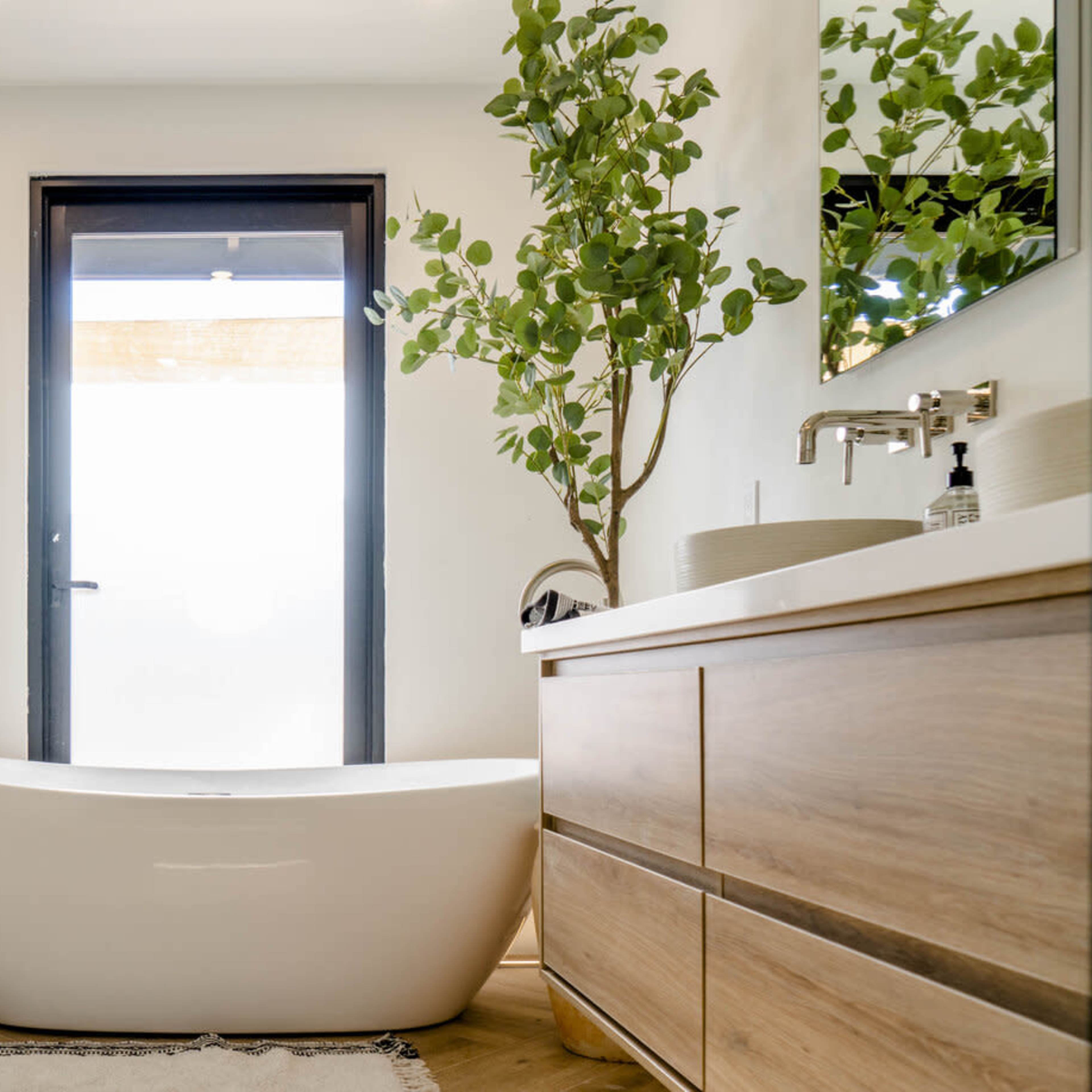 The bathroom features a freestanding white bathtub, a wooden vanity with drawers, and a large window framed in dark wood that opens to an outdoor view.