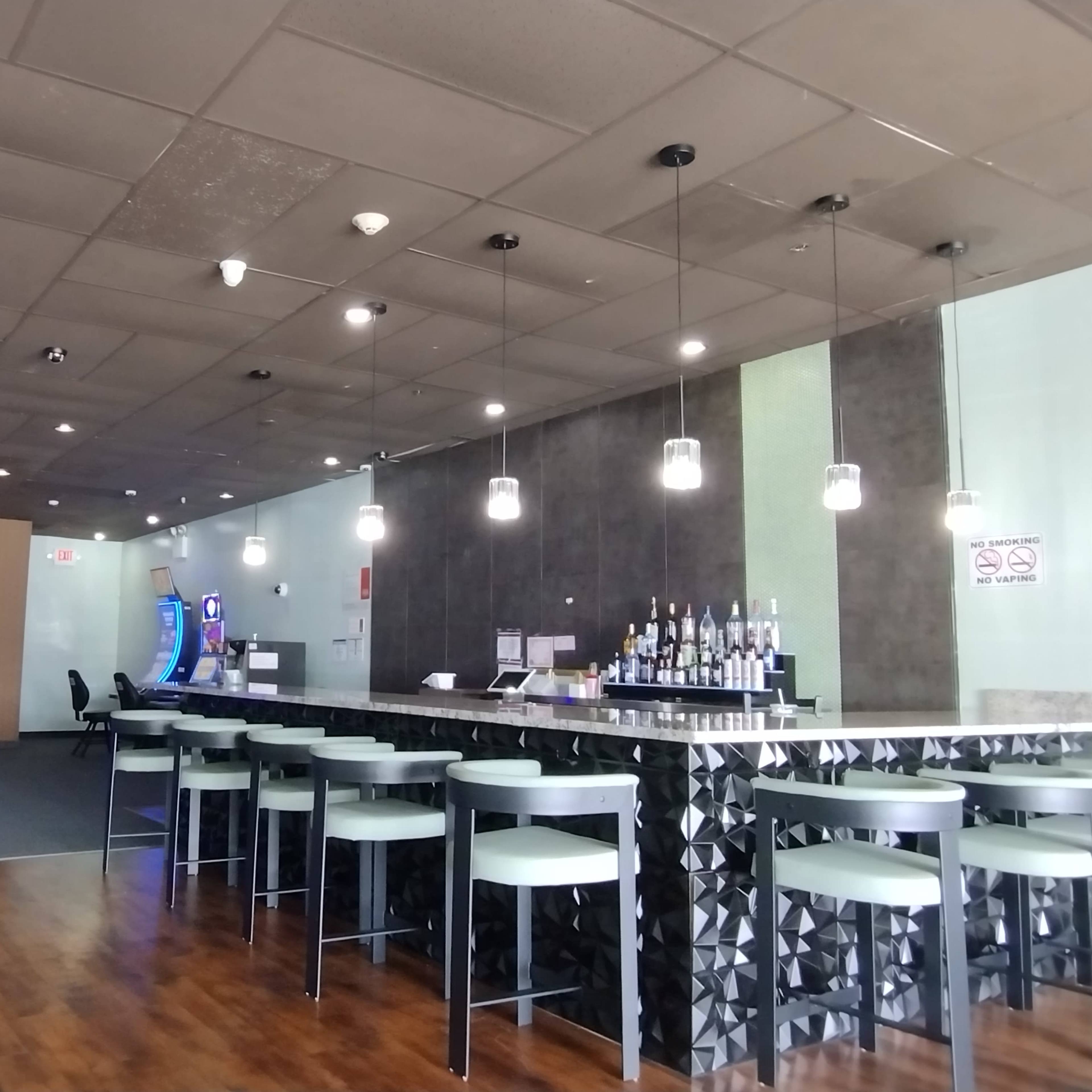 An interior of a modern bar with a counter, pendant lighting, and a row of stools, alongside gaming machines in a lounge area.