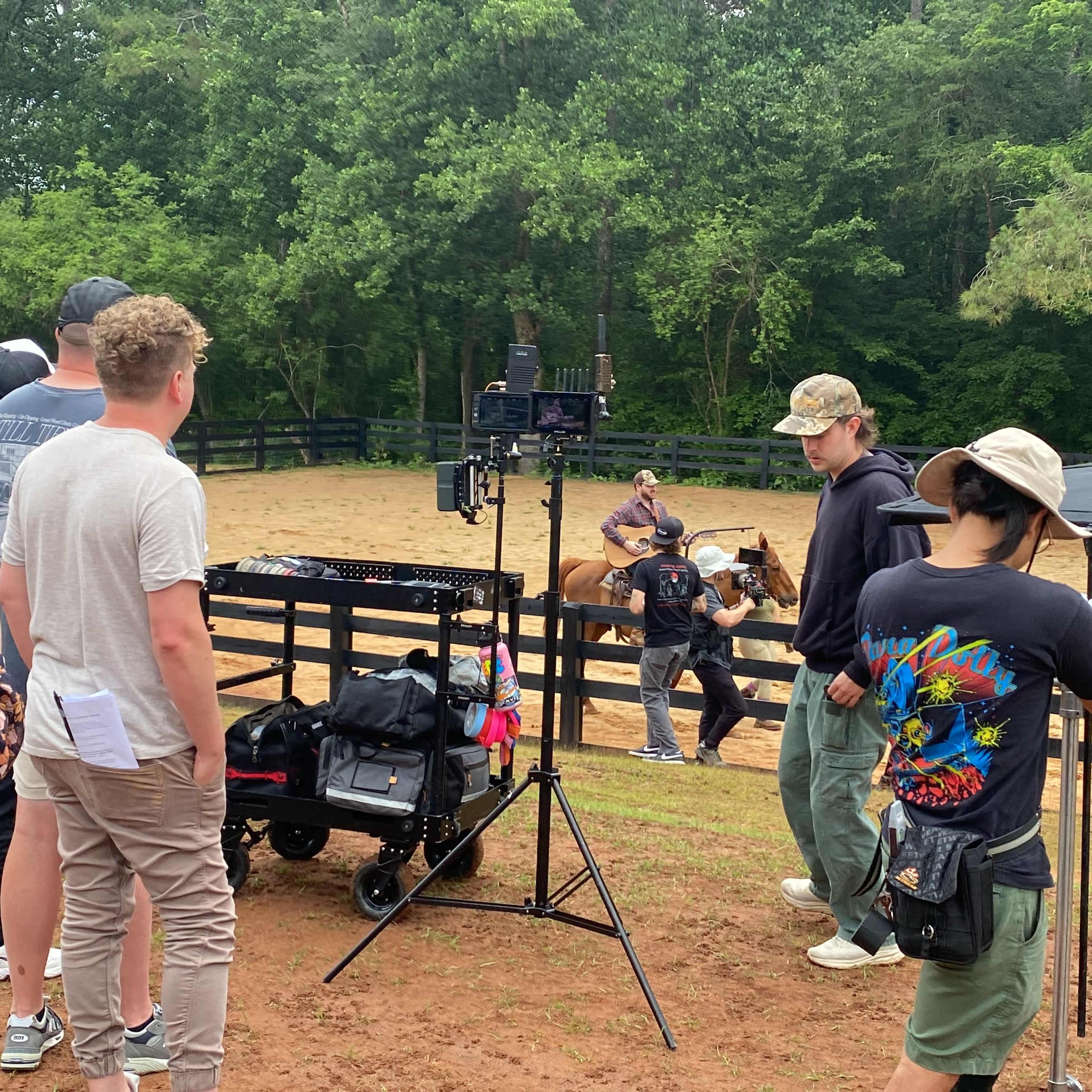 A group of people is gathered near a filming setup in an outdoor area with a horse and a rider in the background.