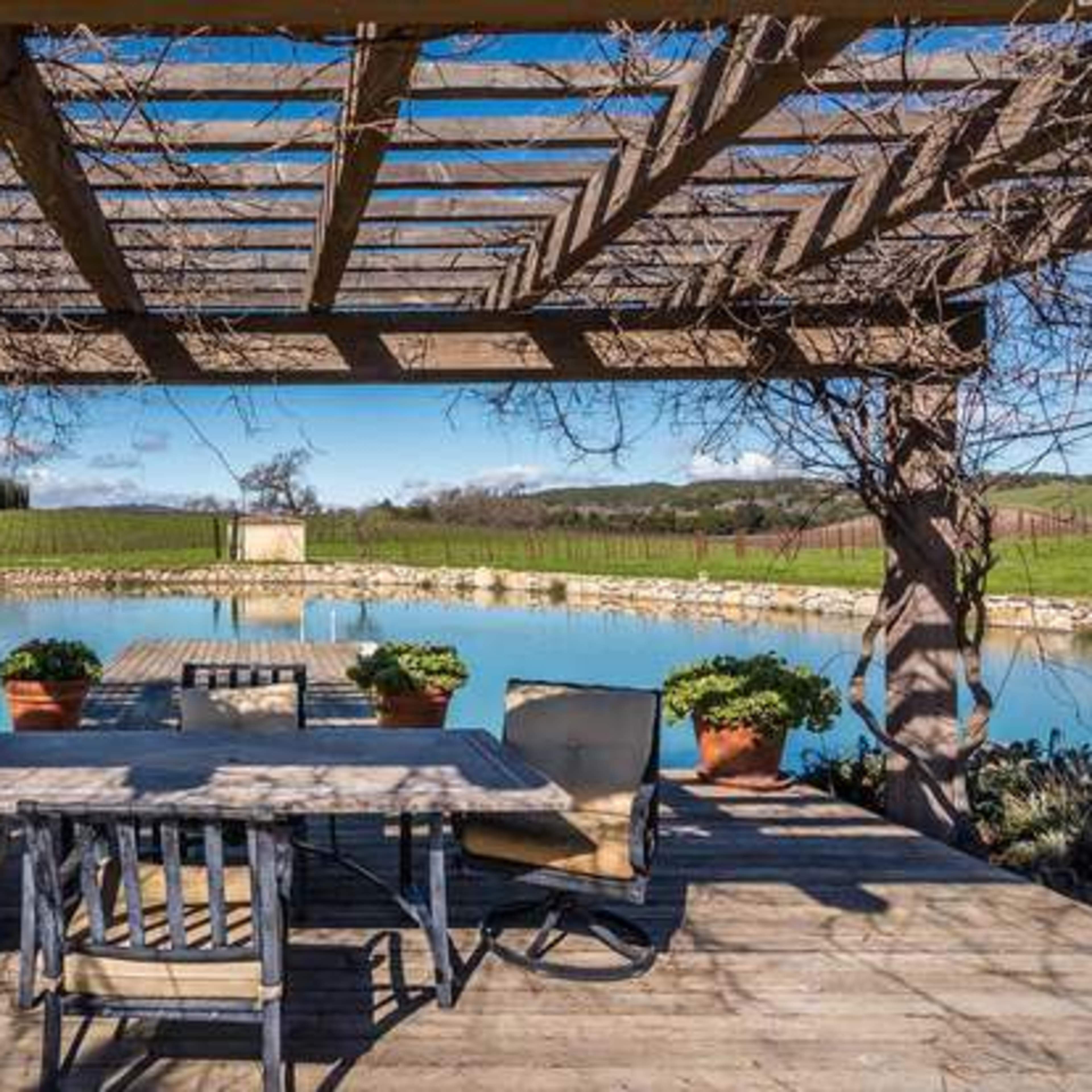 A wooden patio with a table and chairs overlooks a calm blue pond surrounded by greenery.