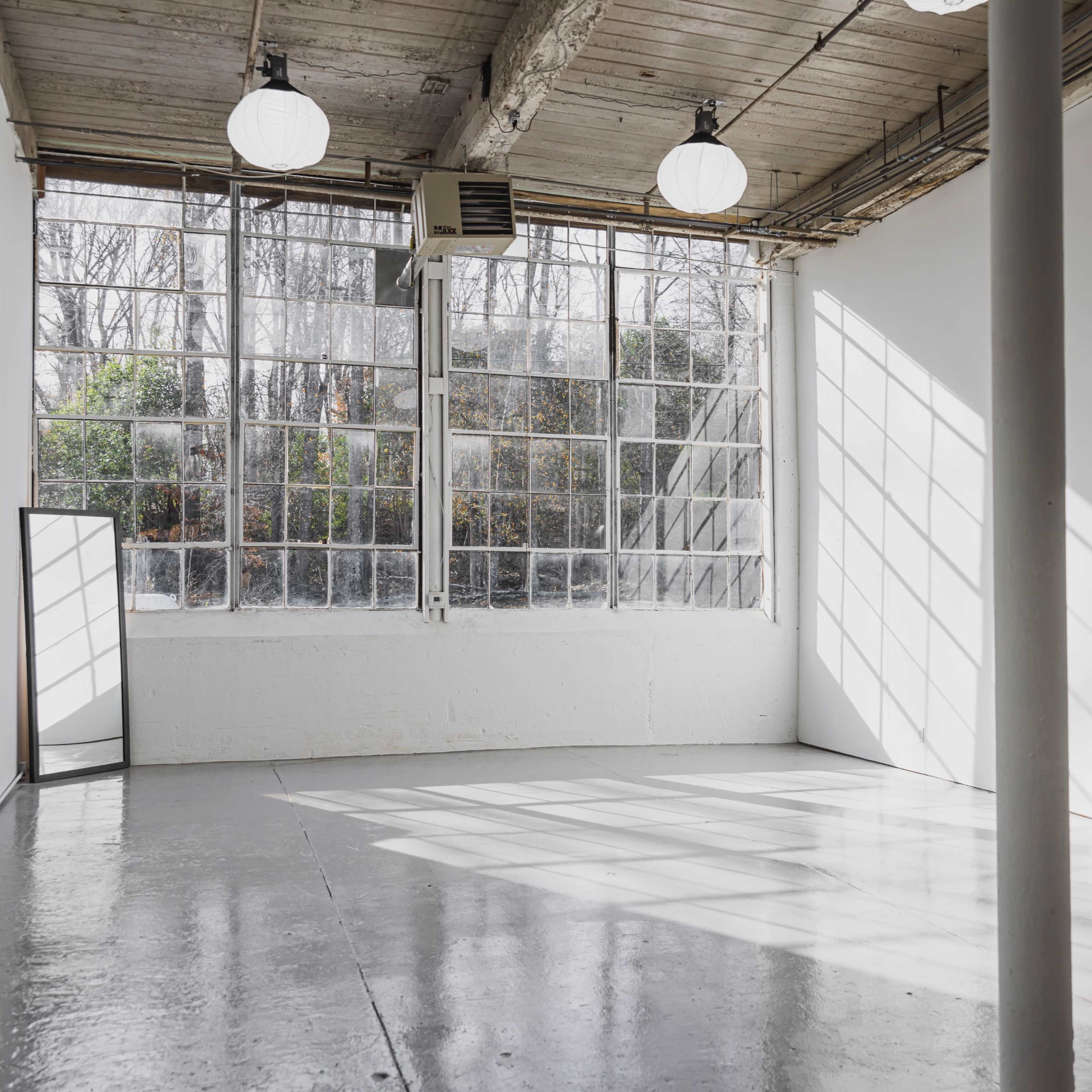 A spacious, well-lit studio with large windows, a mirror, and a concrete floor.